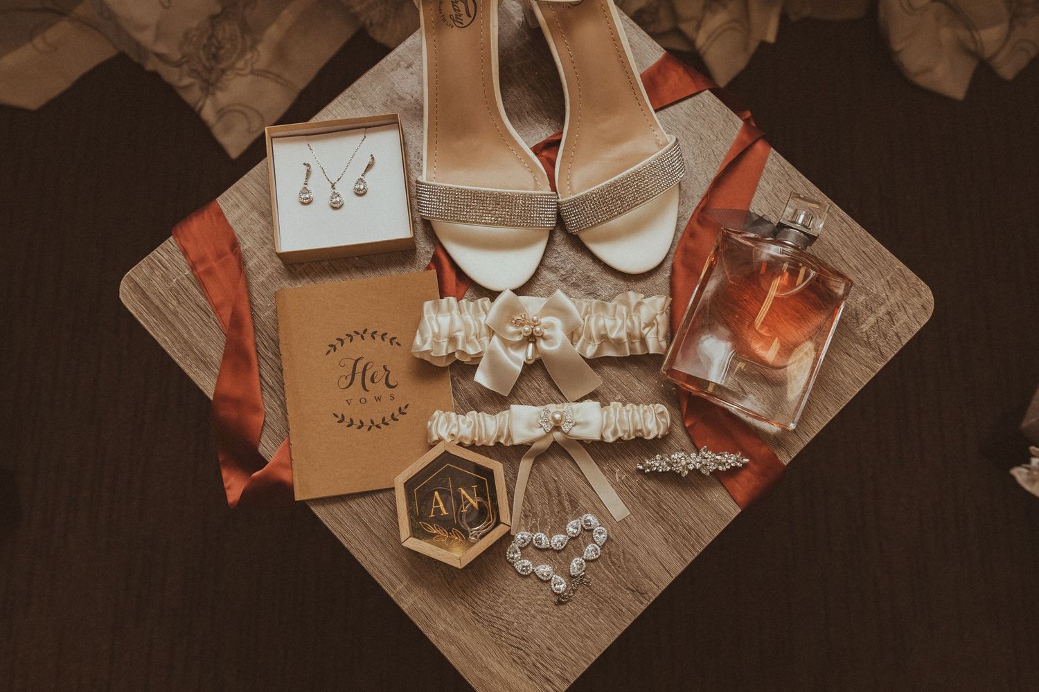 Wedding accessories including white heels, perfume, garters and jewelry arranged on wooden surface.