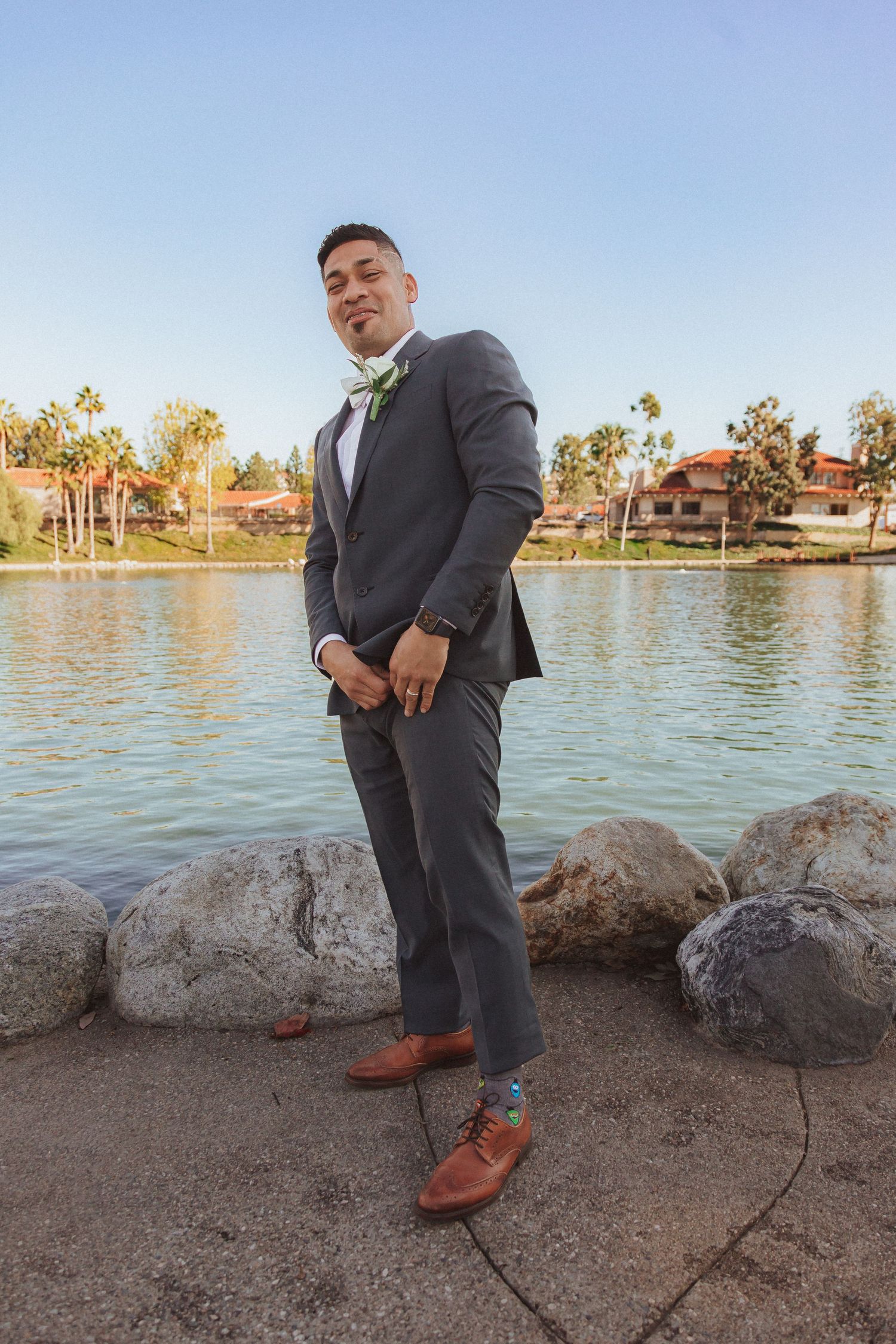Wedding portrait by a lake showing groom in grey suit striking relaxed pose against scenic backdrop.