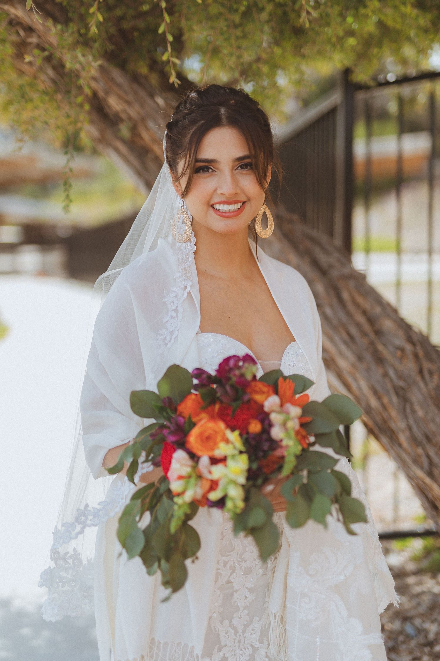 A bride in a lace veil holds a vibrant burgundy and orange bouquet against a sunlit garden backdrop.