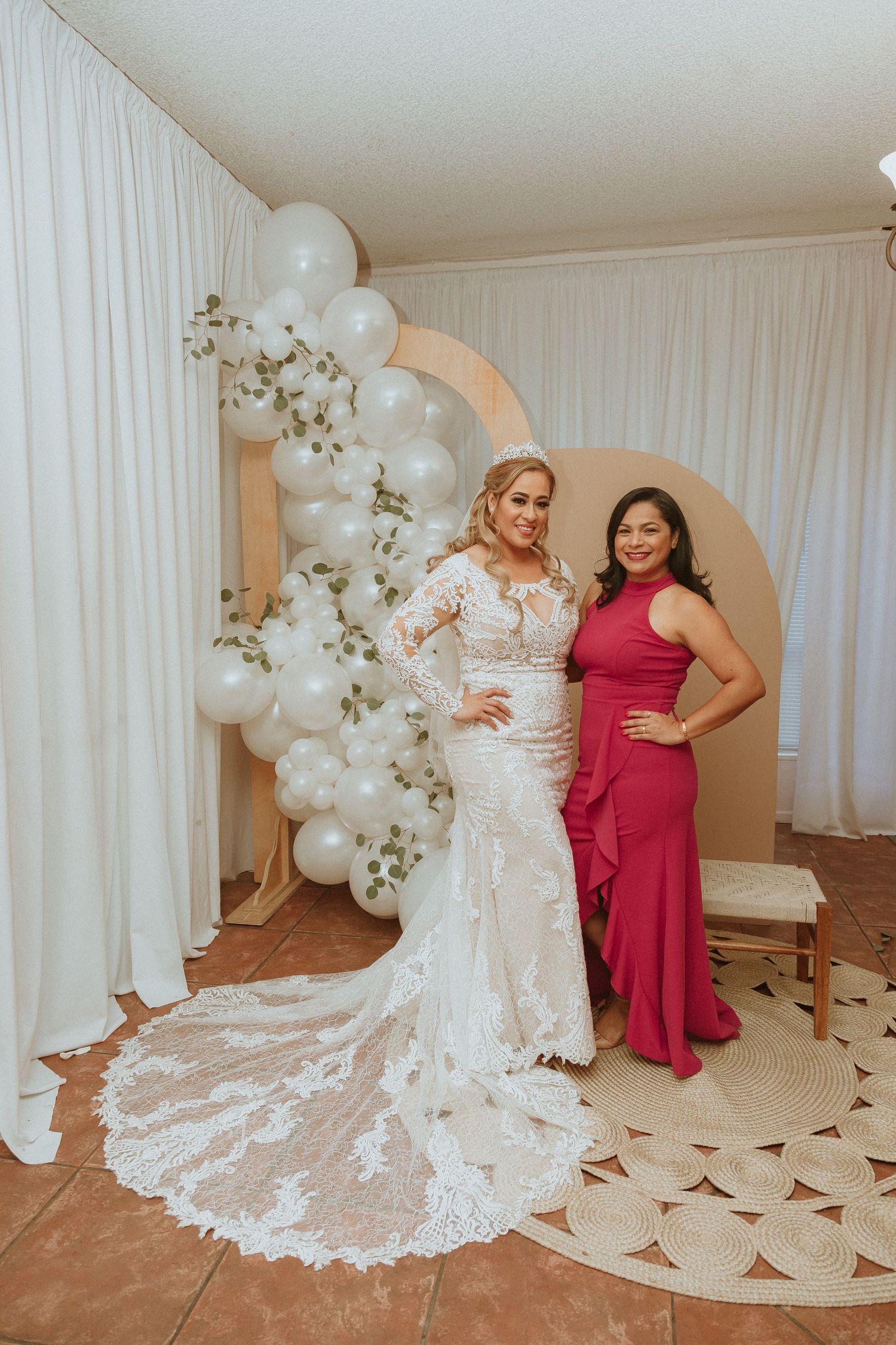 Wedding photo featuring white lace gown and red formal dress against balloon arch backdrop.
