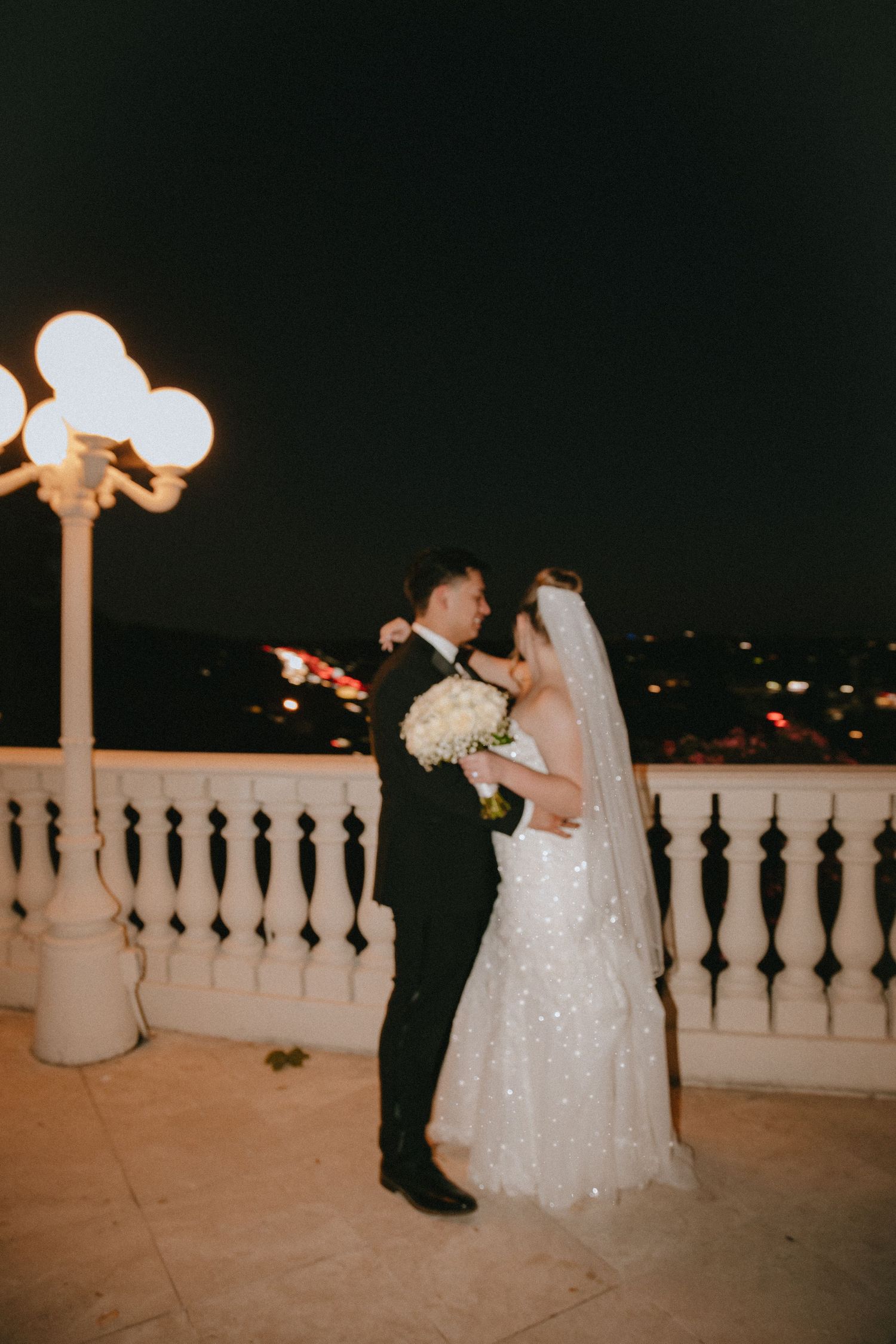 A romantic nighttime wedding moment on a balcony with ornate railing and glowing lamp post.