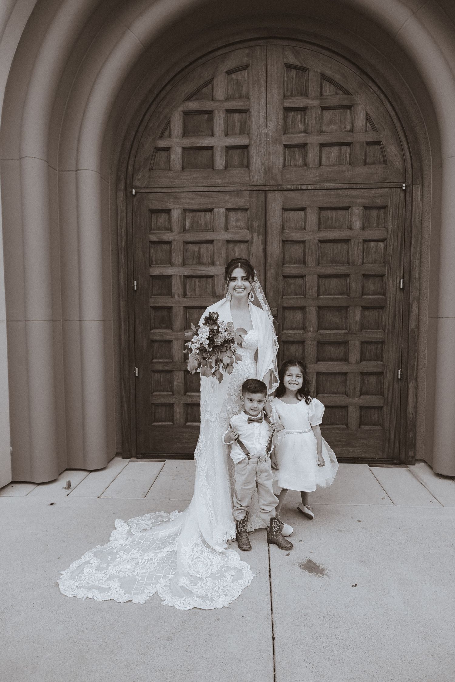 Black and white wedding photo showing bride and children posing before grand wooden doors.