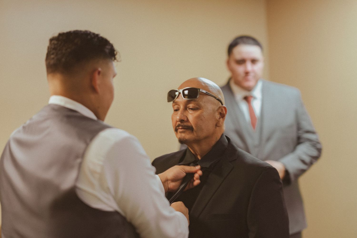 Several people in formal attire assist with tie adjustments in preparation room.