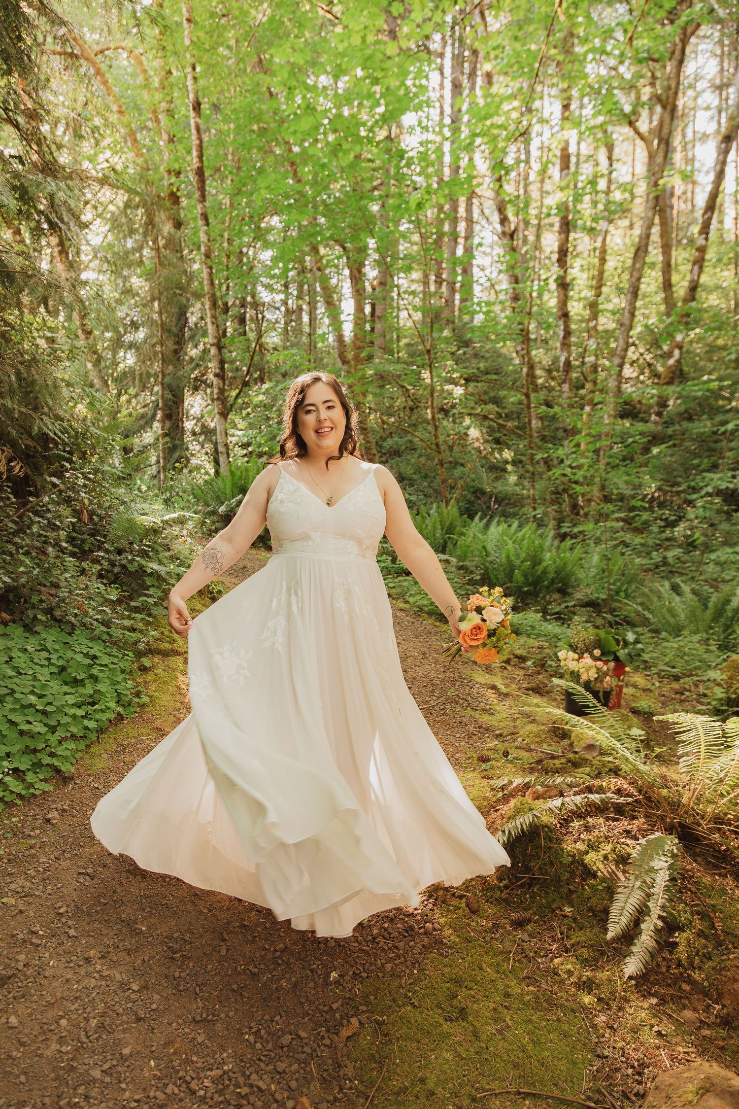 A person in a white wedding gown stands with arms outstretched on a forest trail.