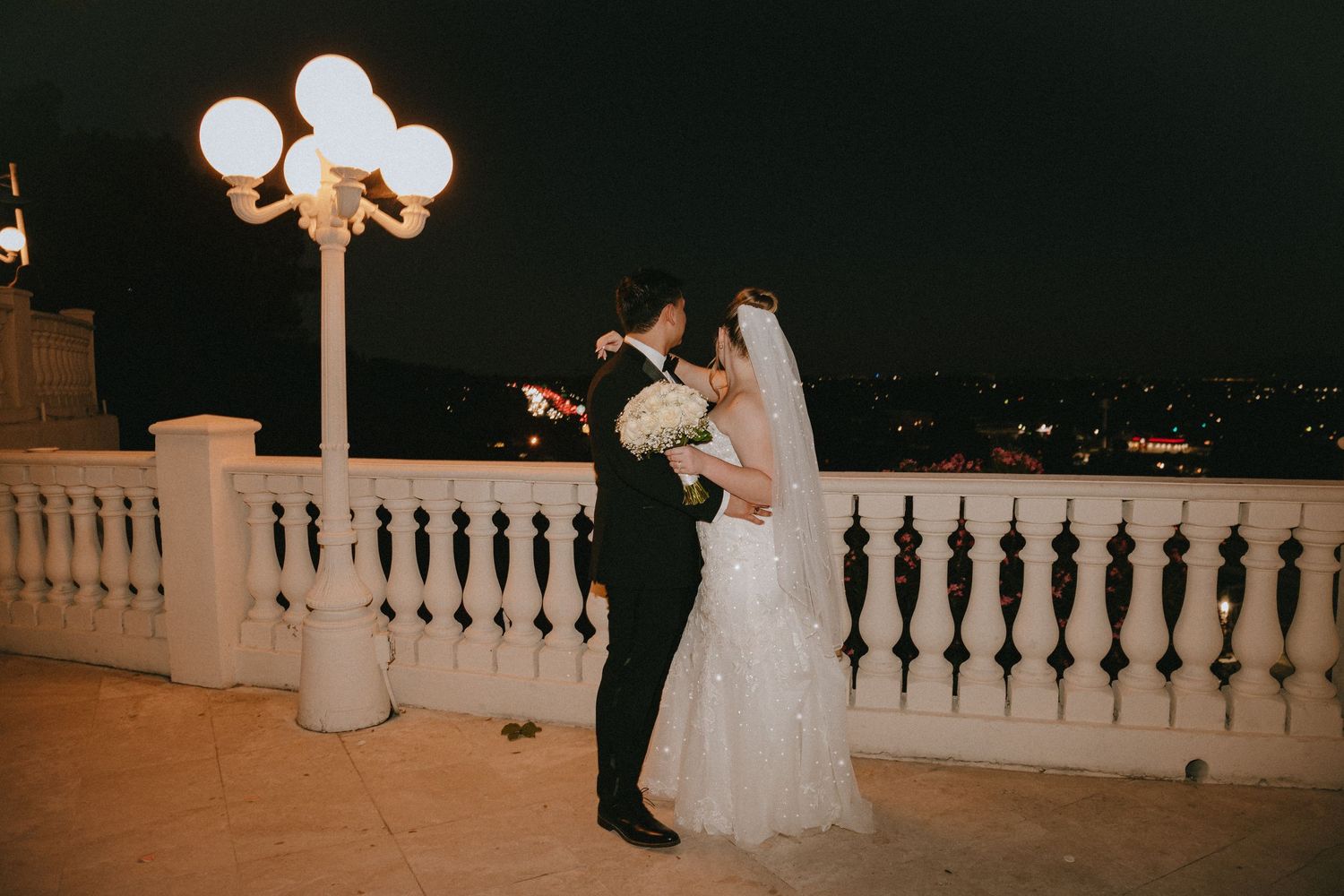 Evening wedding photo showing couple on white balcony with vintage lighting.