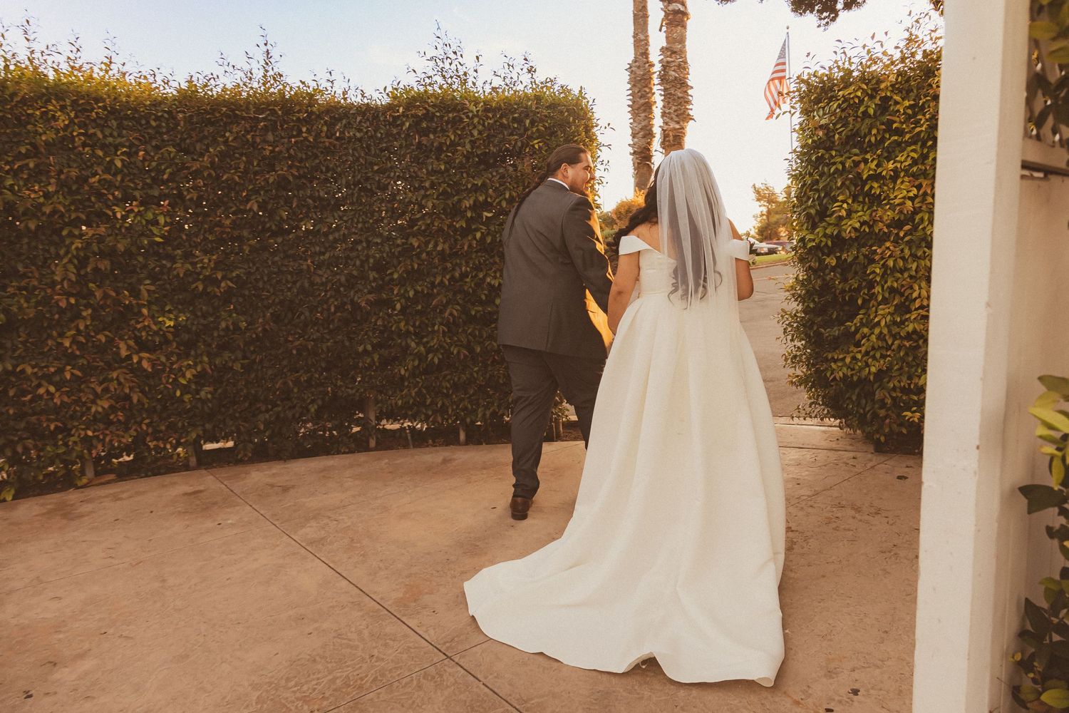 Bride and groom walk along a paved path surrounded by manicured hedges.