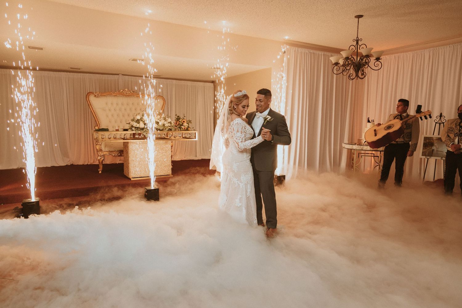 Couple shares first dance on fog-covered dance floor with sparkling cold firework fountains in elegant reception venue.