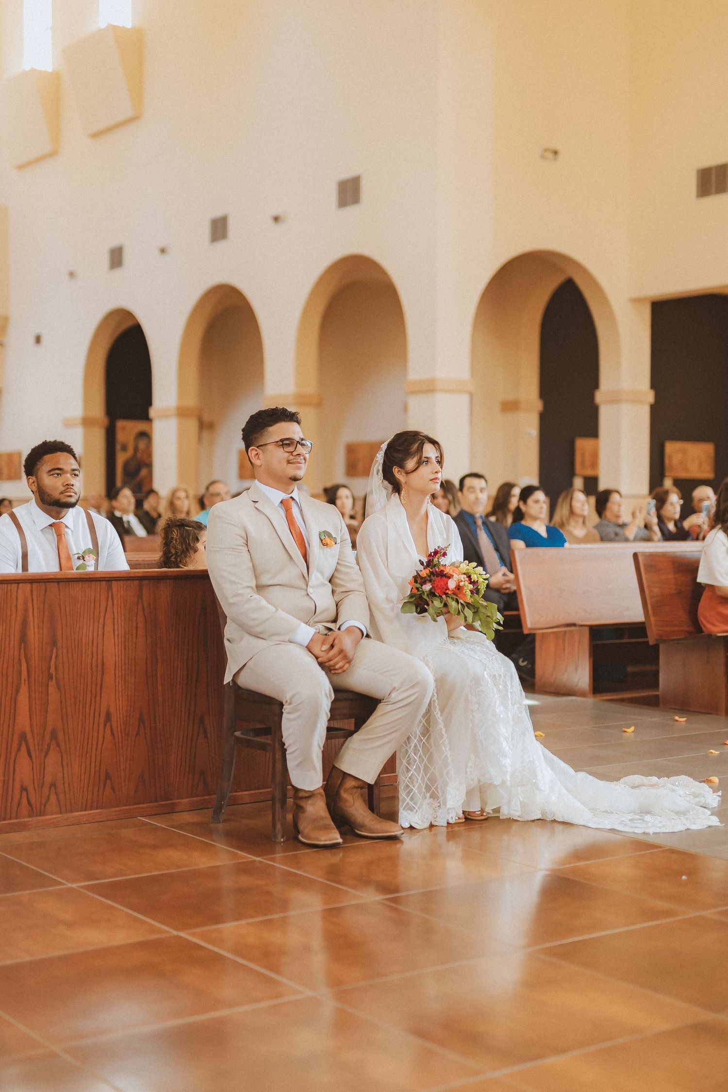 Newlyweds seated in church during wedding ceremony with guests in background.