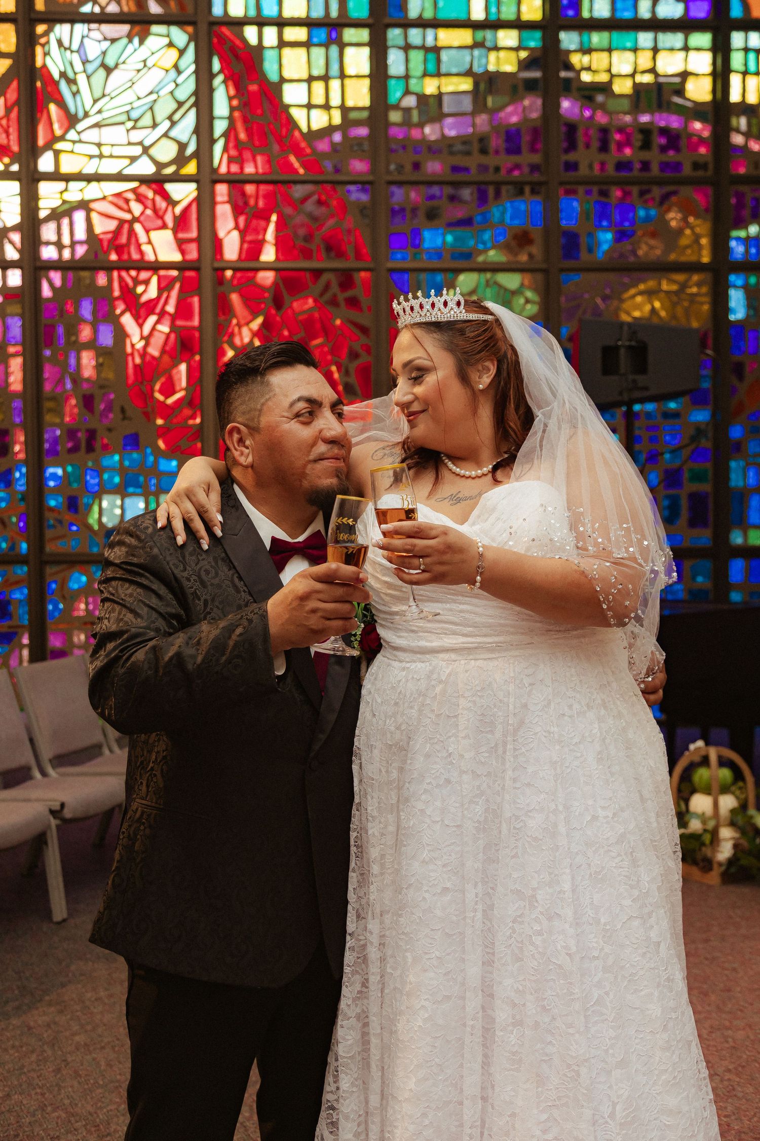Newlyweds share champagne toast in front of colorful stained glass church windows.
