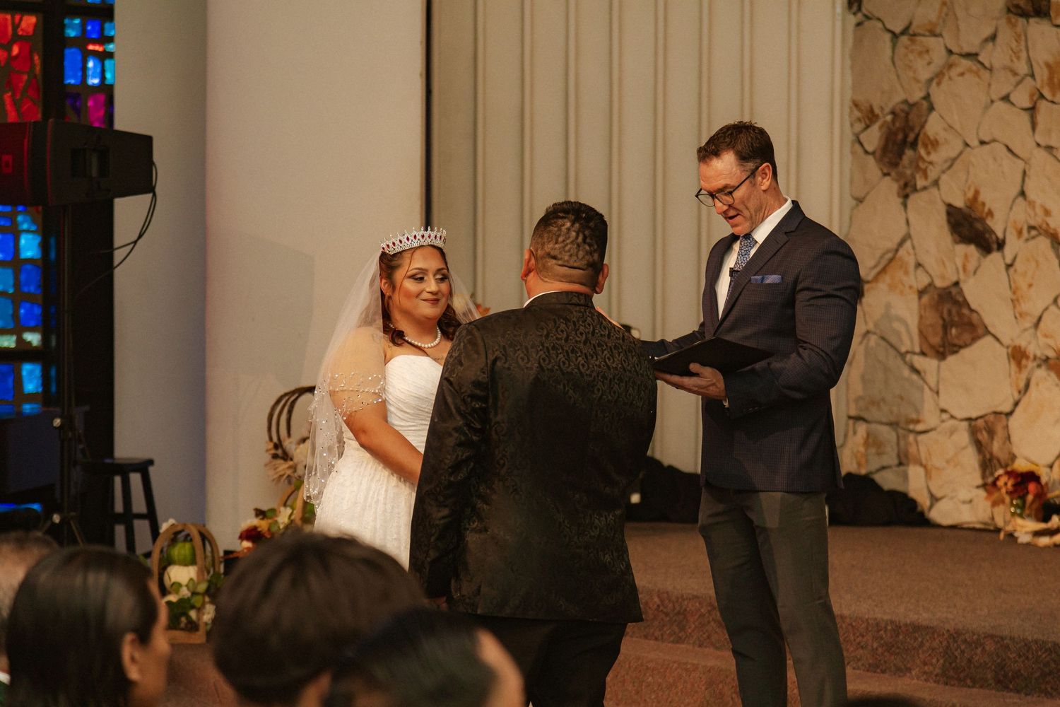 Intimate wedding ceremony takes place in front of stone wall with stained glass windows in background.