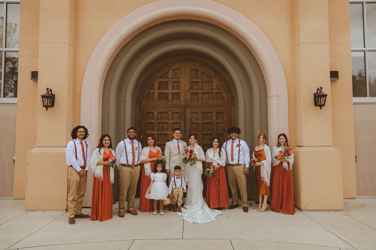 Wedding group photo with bridal party wearing coral and khaki colors against church architecture.