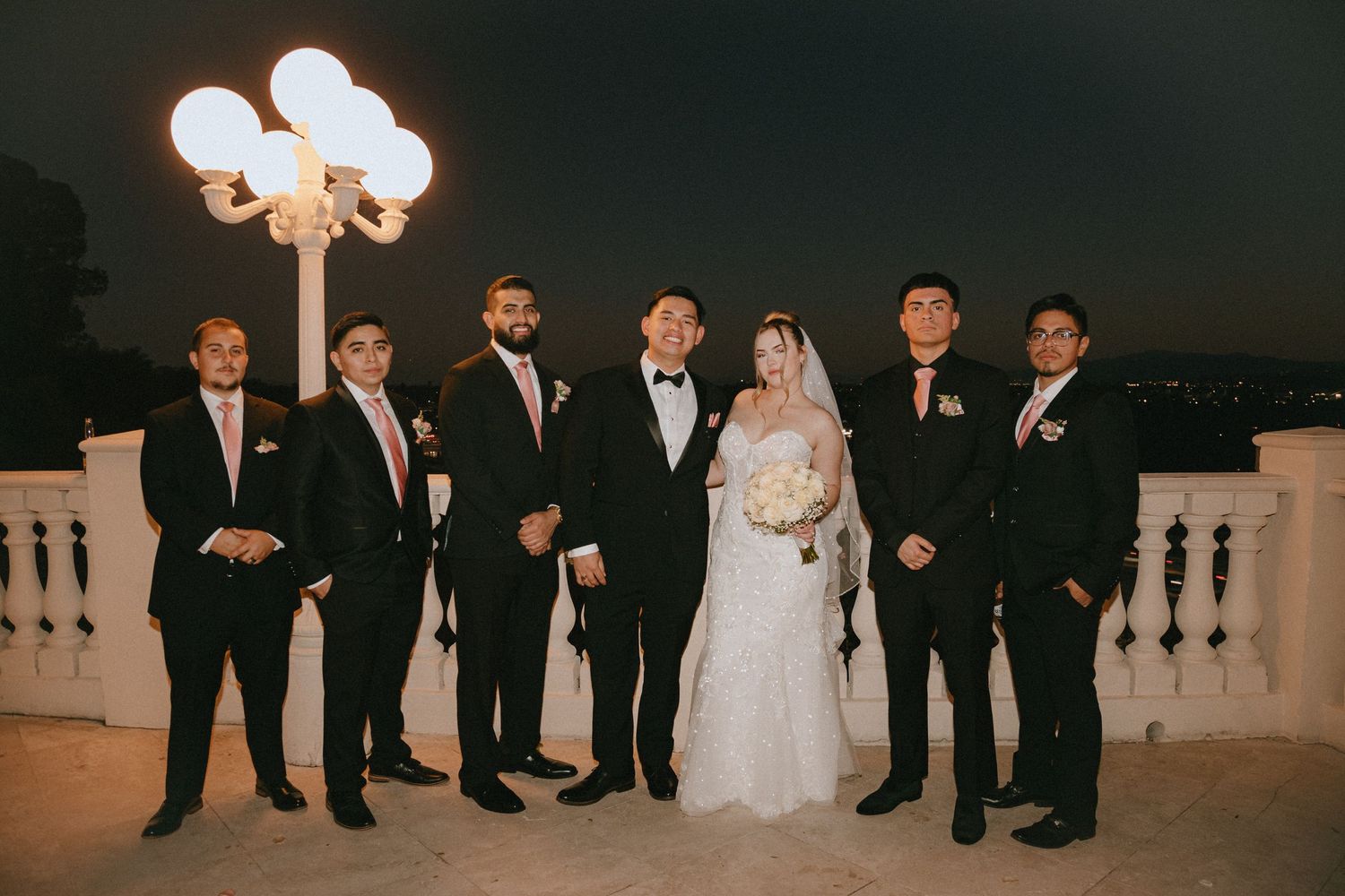 Wedding party in formal attire poses under ornate lamp post at dusk.