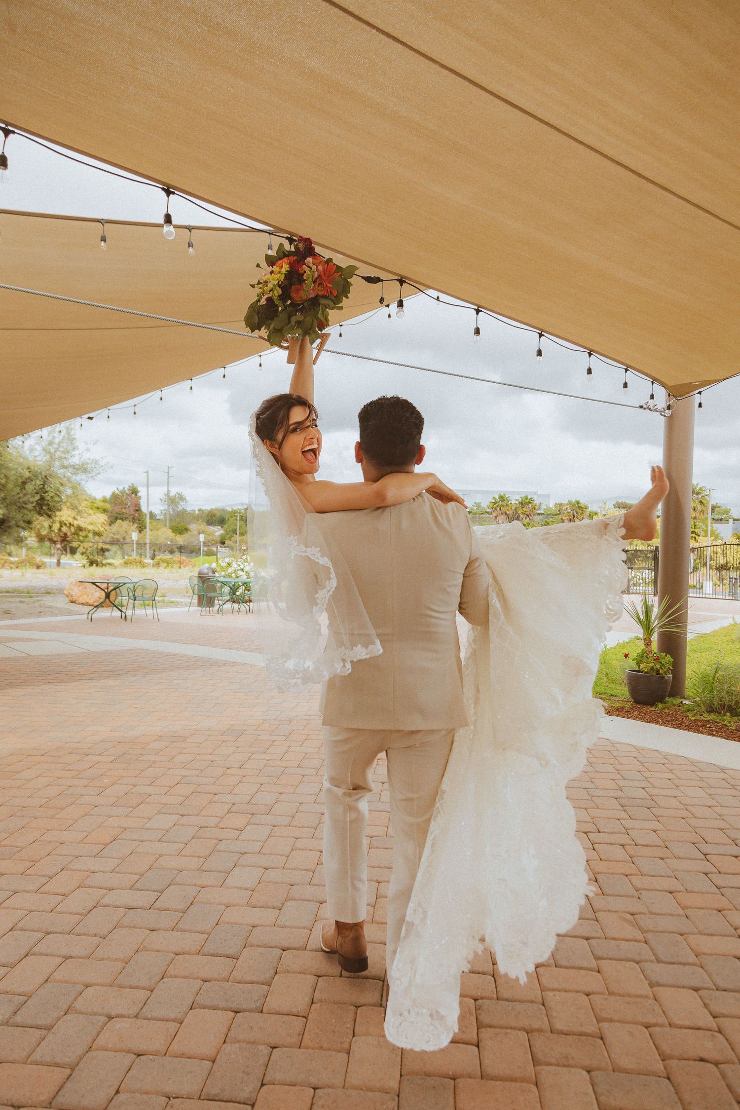 Newlyweds dance together under a beige canopy with string lights and flowing white dress fabric on a brick patio.