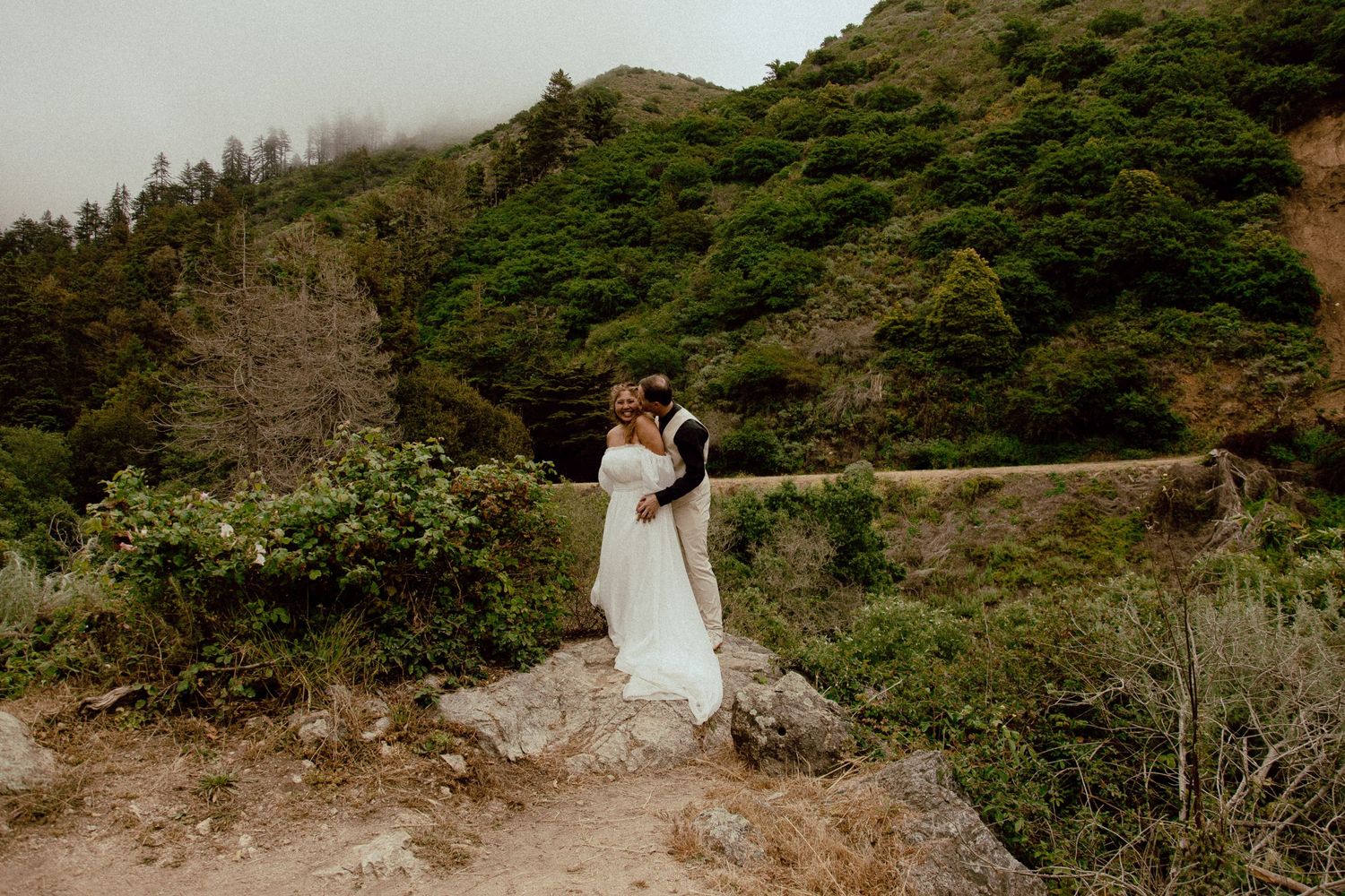 A couple in wedding attire embraces on a misty coastal clifftop surrounded by green hills.