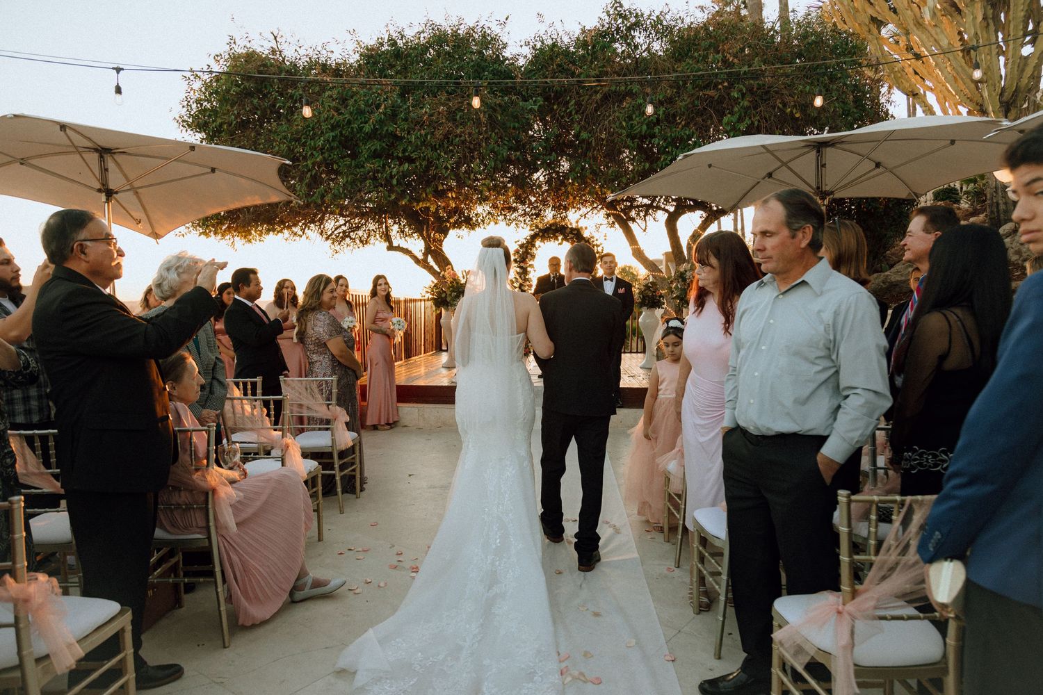Wedding guests line the aisle during an outdoor ceremony with market lights and trees.