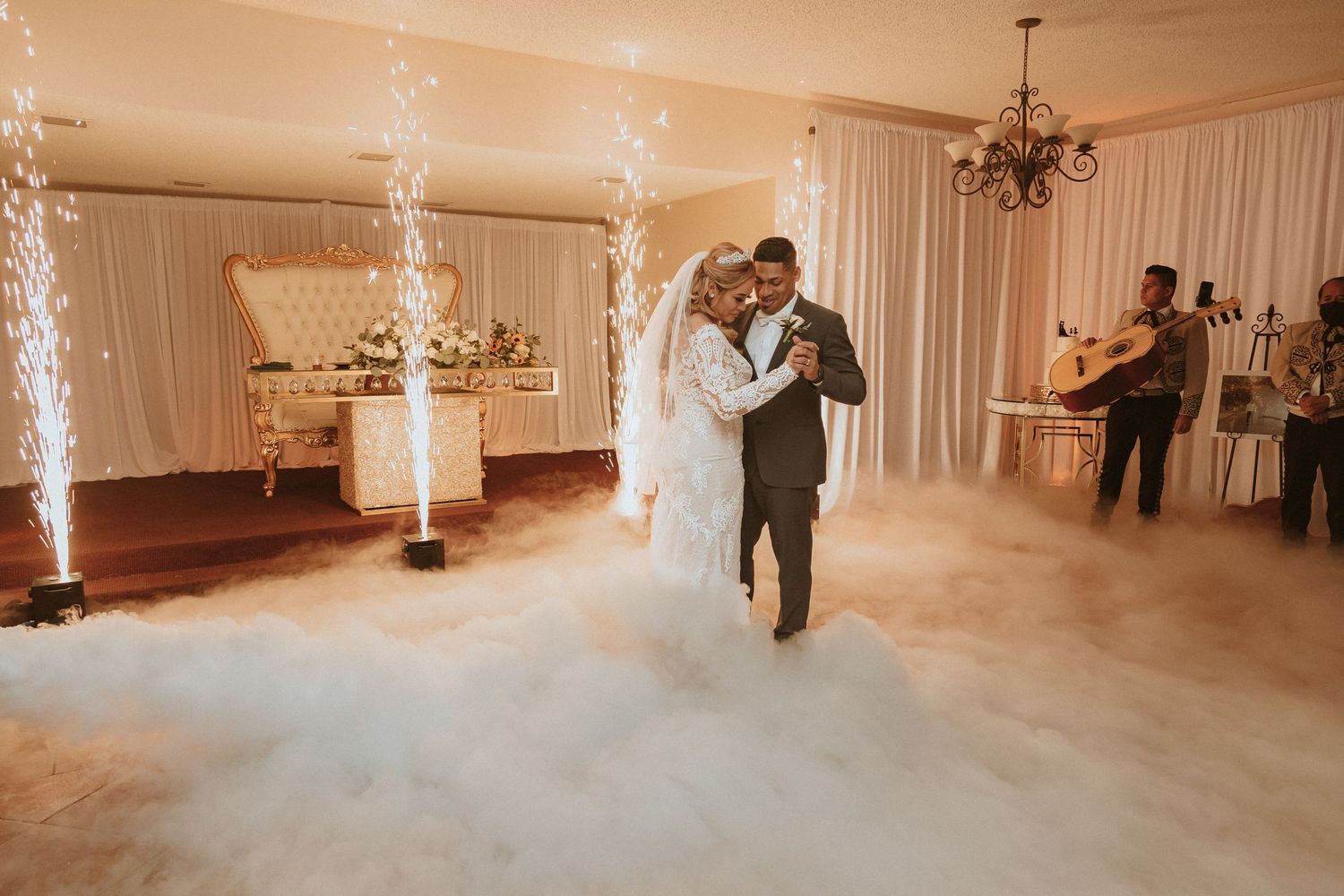 Wedding couple dances through atmospheric fog while sparkler fountains illuminate elegant draped reception space.