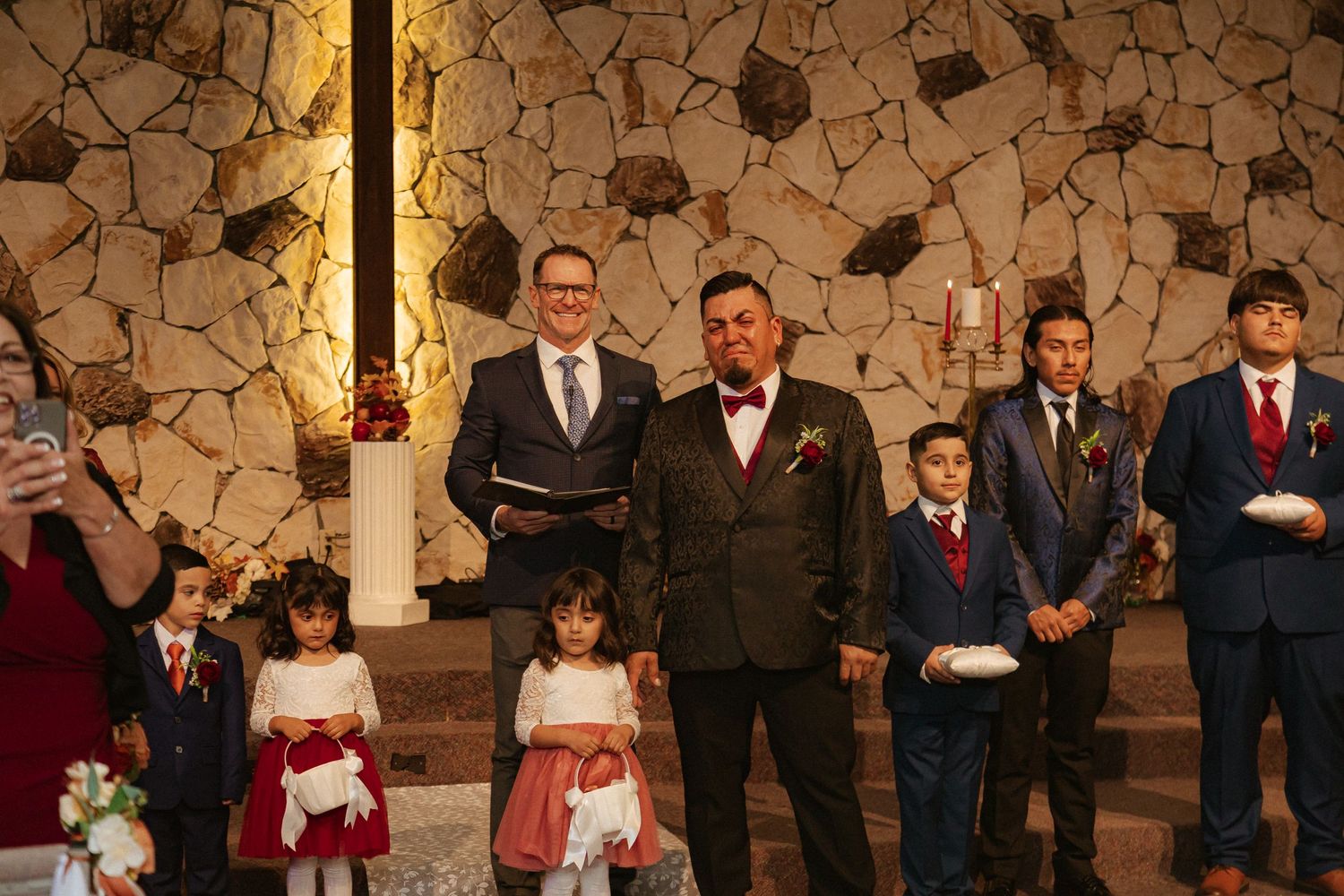 Wedding ceremony participants gather at stone-walled altar with flower girls and attendants during church service.