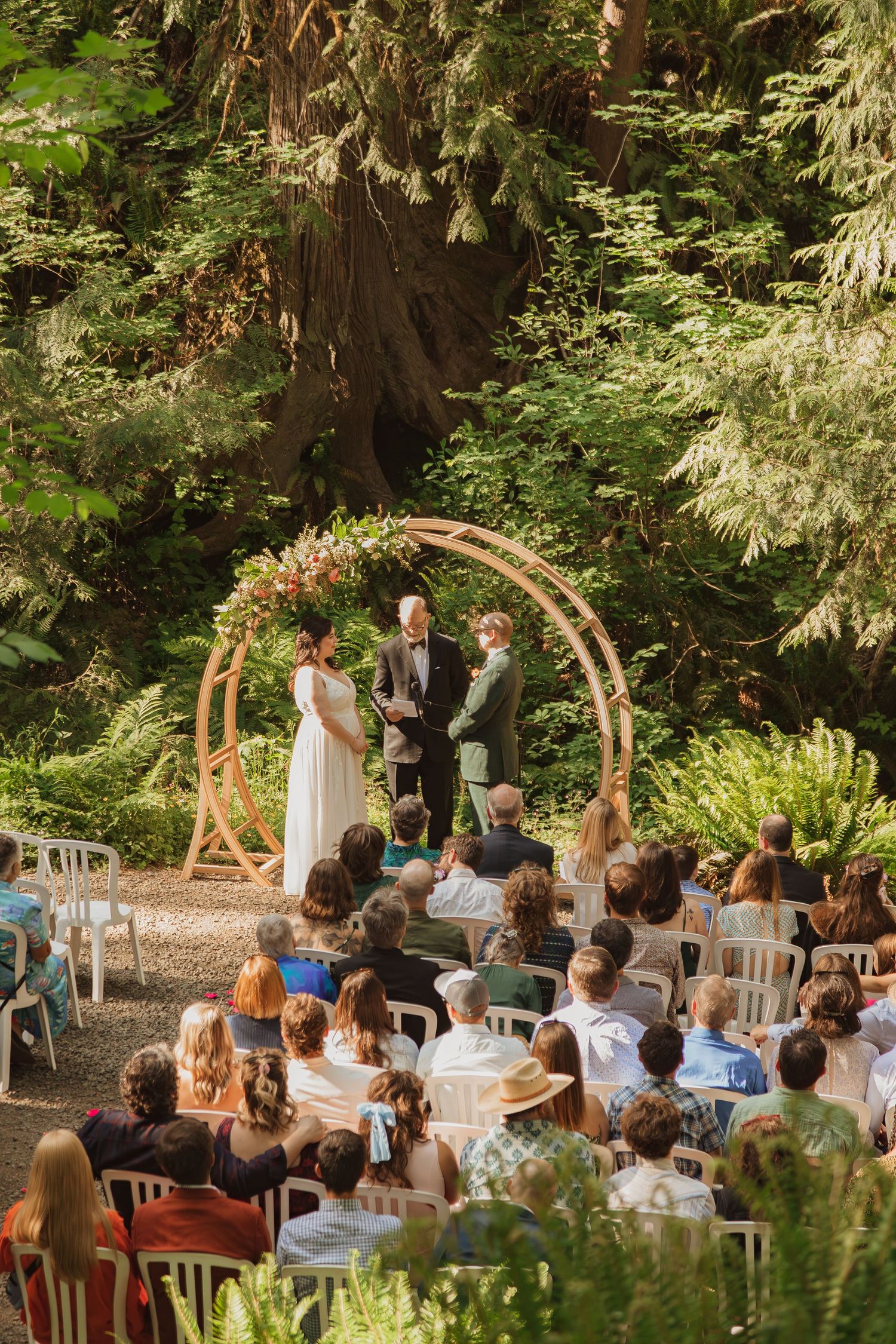 An intimate forest wedding ceremony under a wooden circular arch surrounded by lush greenery and seated guests.