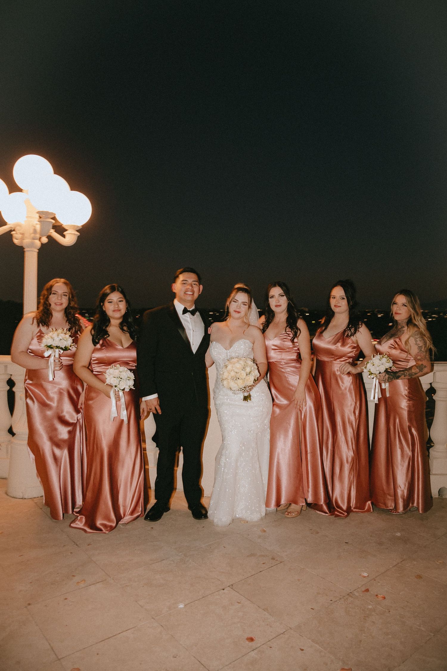 Wedding group photo featuring bridesmaids in matching rose gold gowns on an elegant terrace.