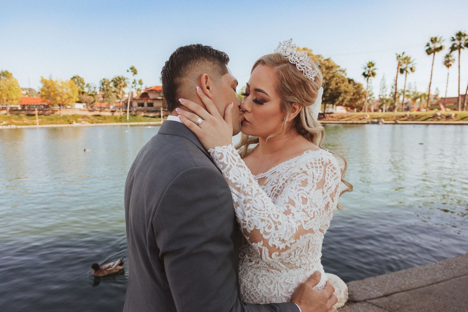 Romantic wedding couple embraces near a tranquil lake with palm trees in the background.