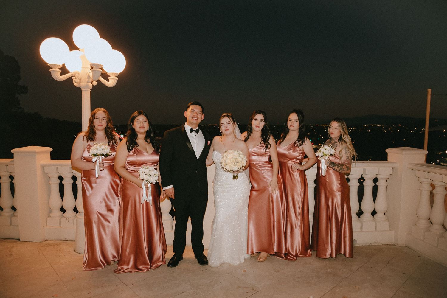 Bridal party in white and rose gold dresses pose on balcony with decorative lighting at night.