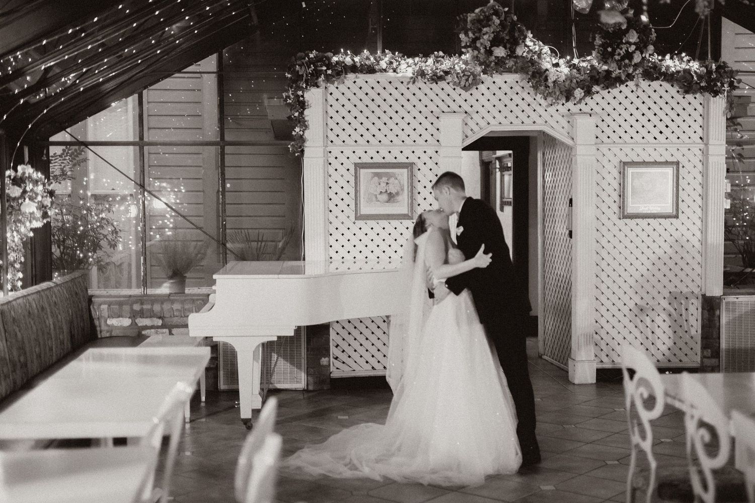 Monochrome wedding photo showing couple embracing by white lattice arch and piano in rustic venue.