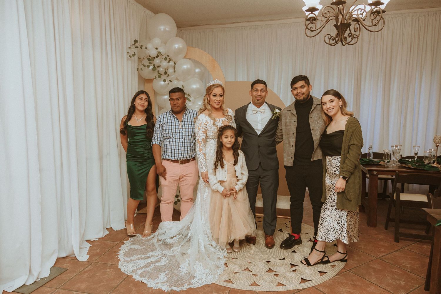 Wedding group photo with family members under elegant balloon arch and chandelier.