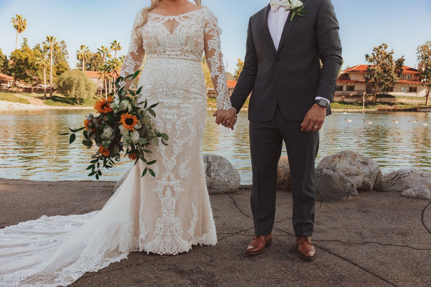 Bride in lace gown and groom in suit hold hands by a lakeside setting.