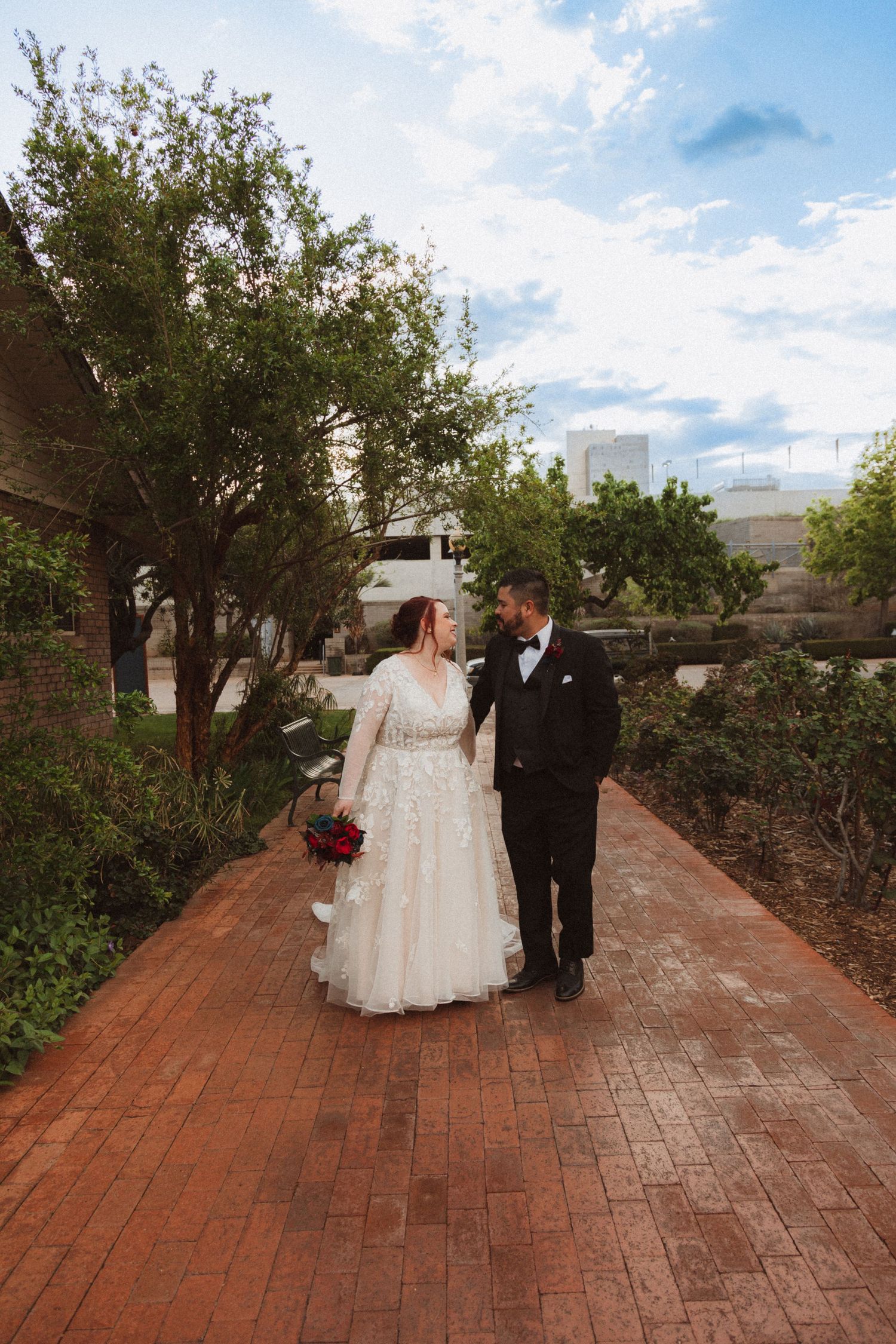 A newlywed couple walks down a brick pathway surrounded by trees and modern buildings.
