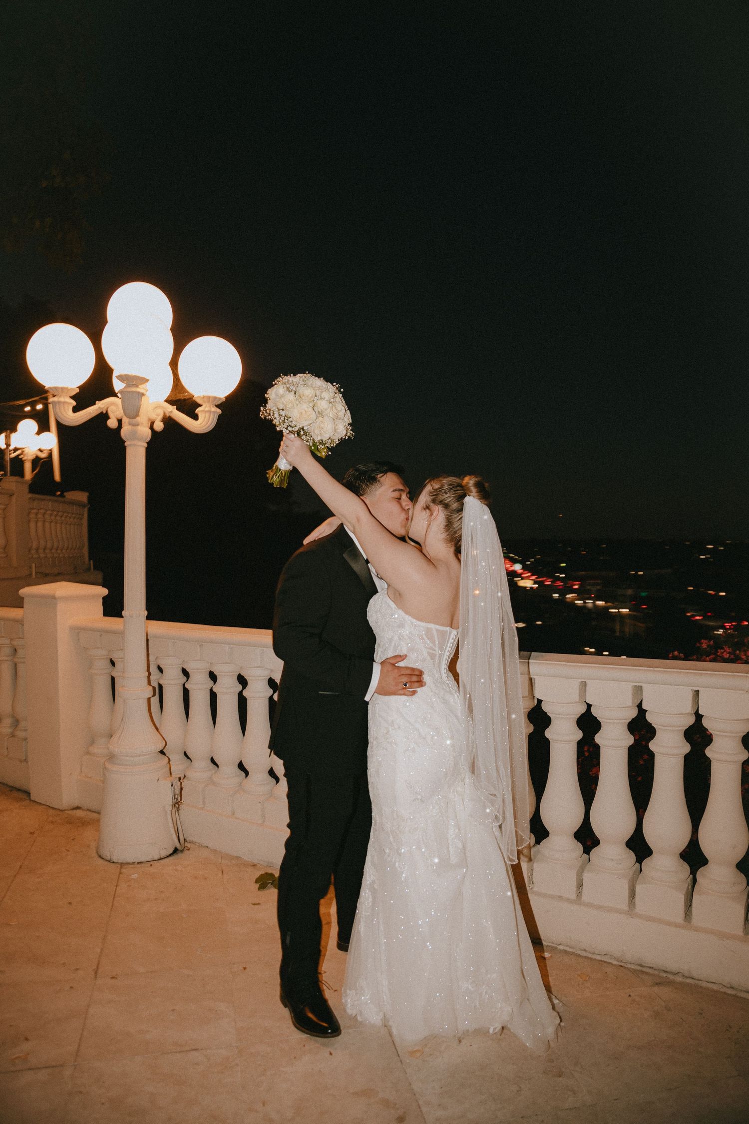 Romantic nighttime wedding photo on a balcony with ornate white railing and glowing street lamps.