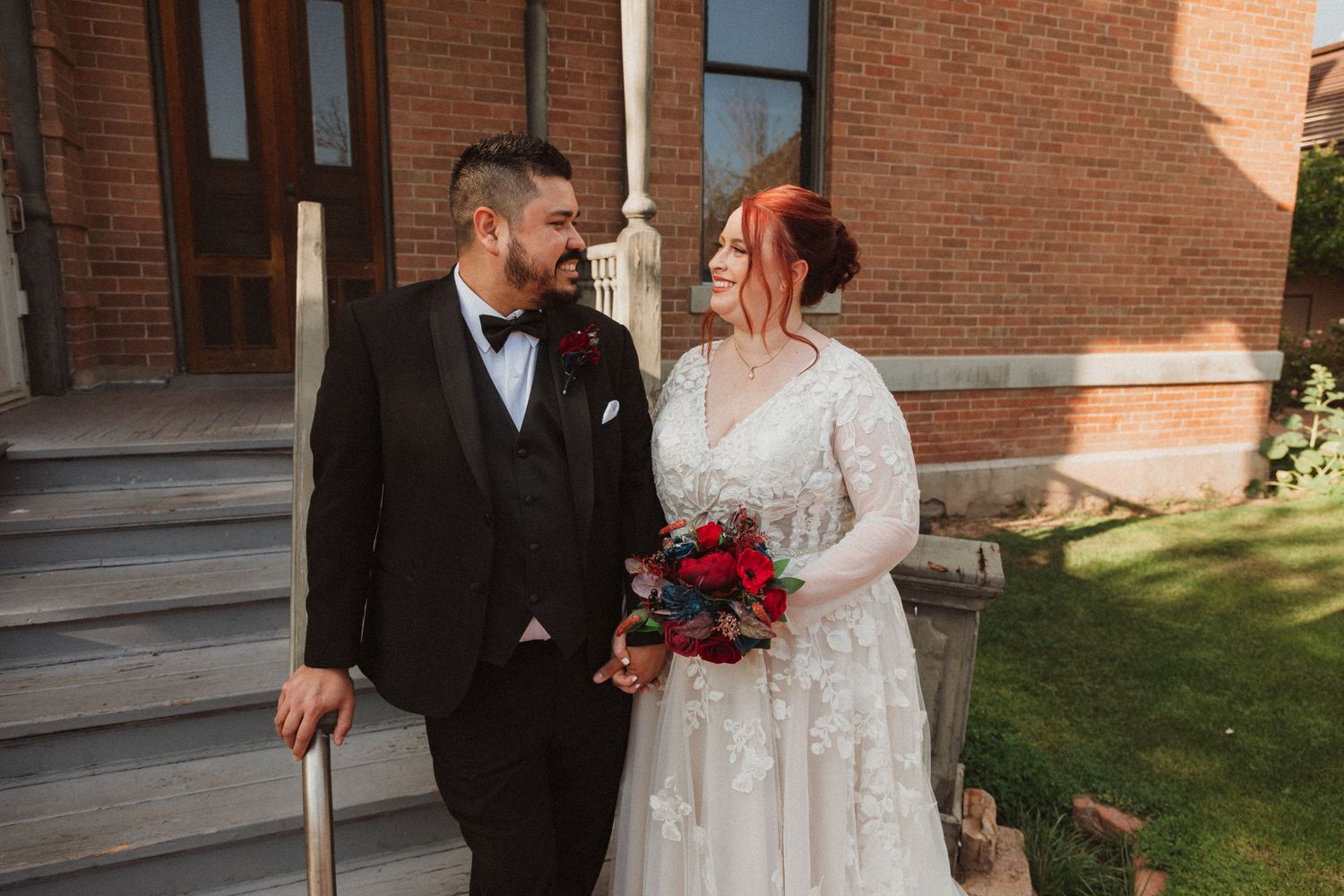 A wedding couple poses on brick steps in front of a vintage building holding a red bouquet.