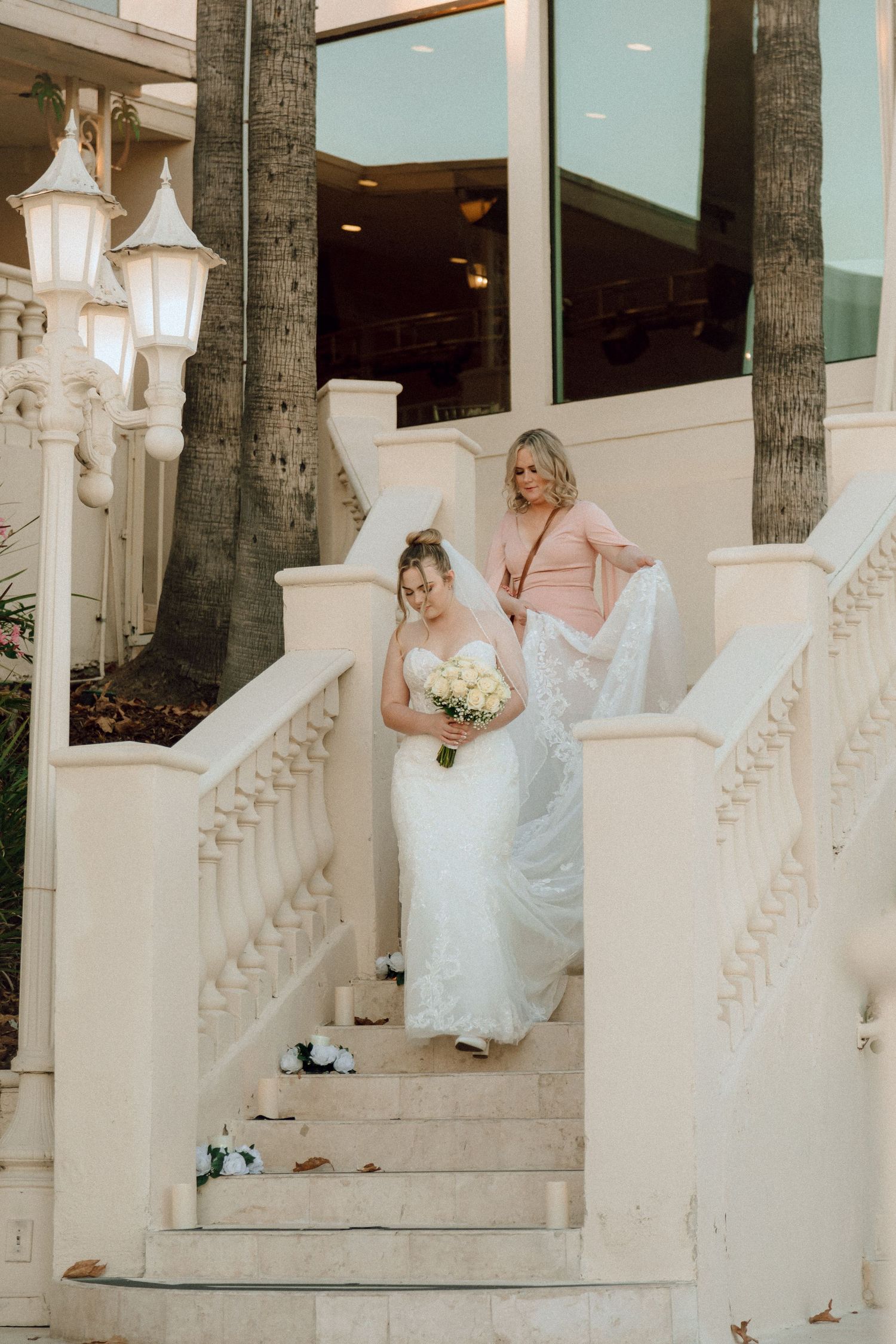 A wedding party descends elegant white stairs adorned with ornate lanterns and palm trees.