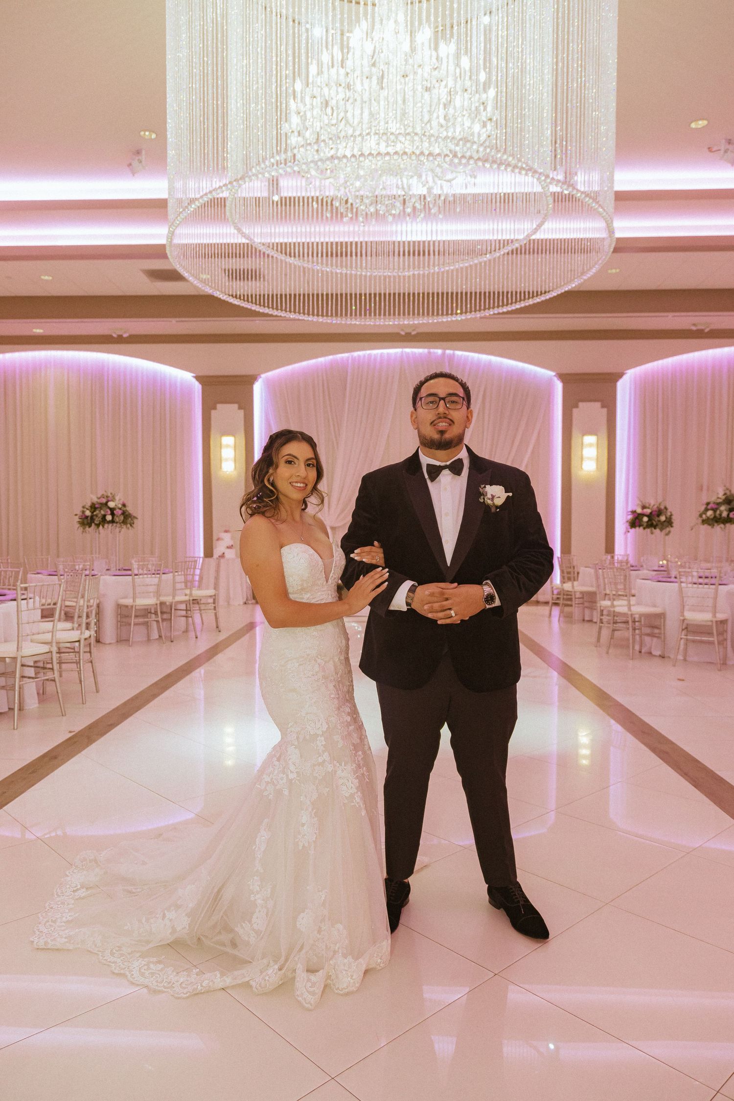 A newly married couple poses elegantly in a luxurious ballroom with pink lighting and crystal chandelier.