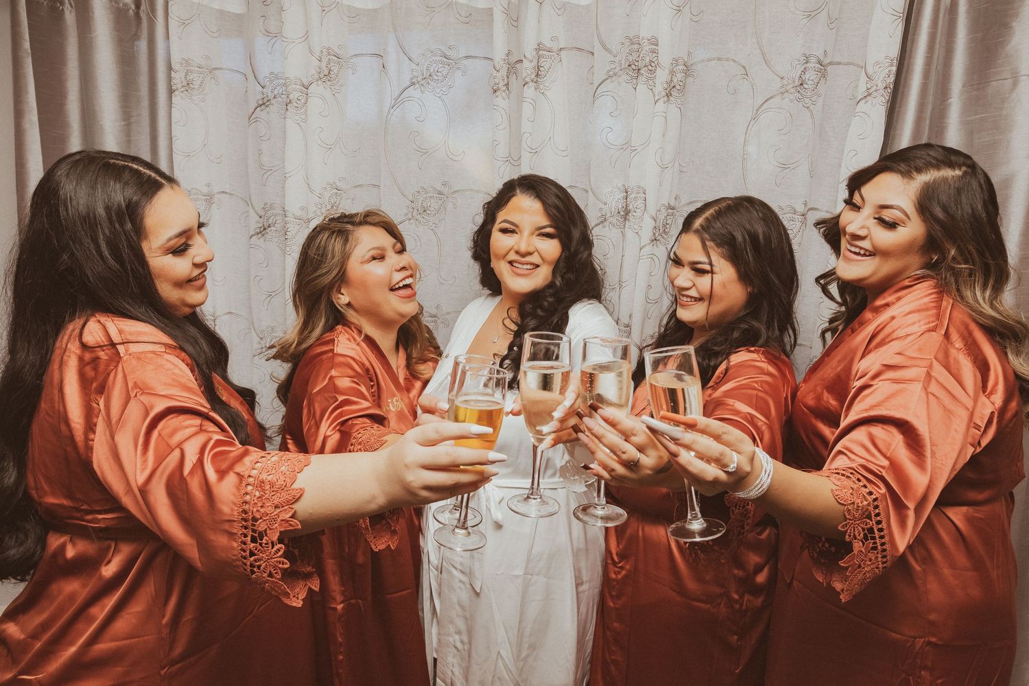 Bridesmaids in coral robes laugh while toasting with the bride in white.