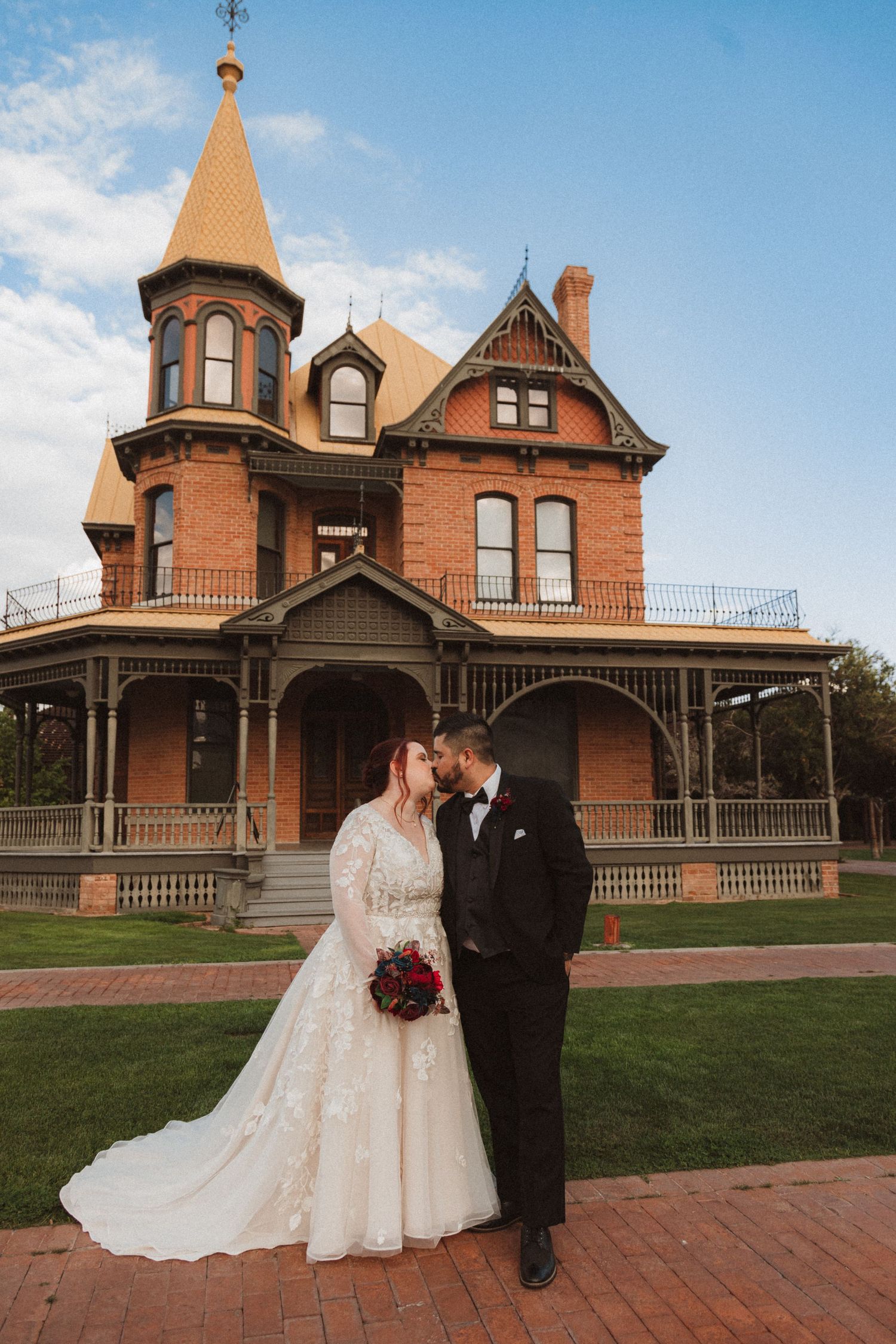 A romantic wedding photo in front of an ornate Victorian house with distinctive tower and wrap-around porch.