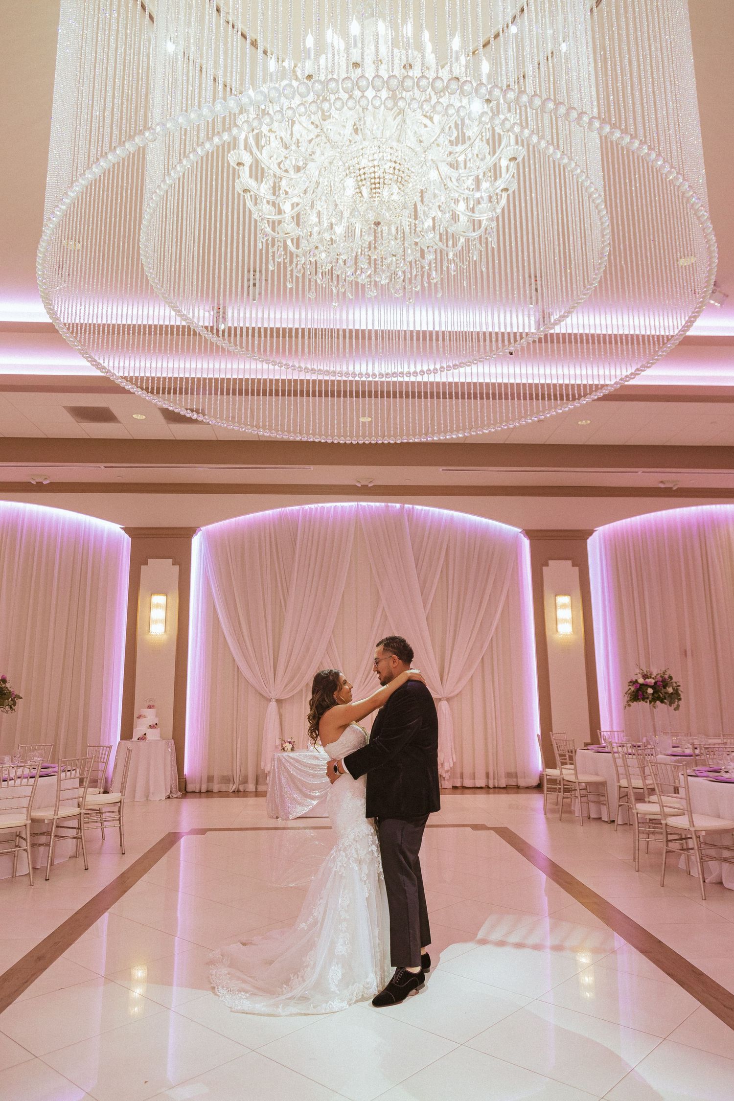 Wedding couple embracing under a magnificent crystal chandelier in a pink-illuminated ballroom.