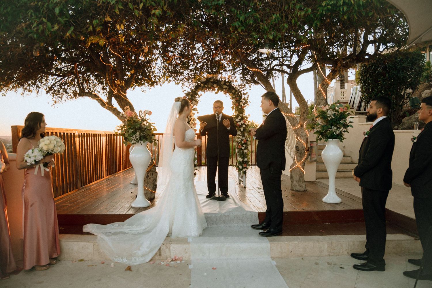An intimate outdoor wedding ceremony with white floral decorations and string lights at sunset.