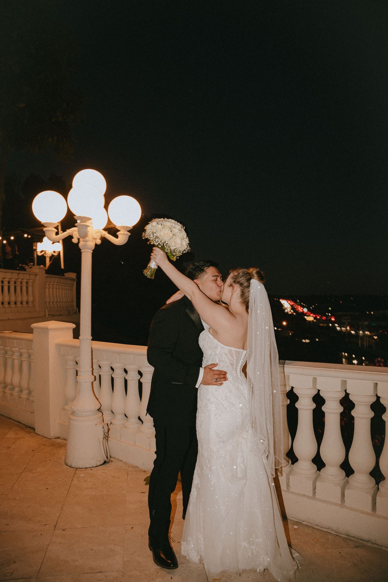 Couple in formal wedding attire embracing on an elegant terrace at night.