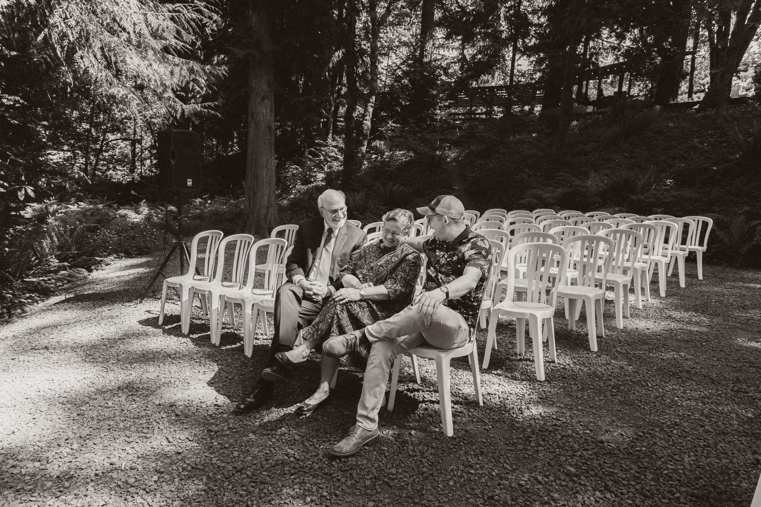 Black and white photo of wedding guests seated in white chairs at an outdoor forest ceremony.