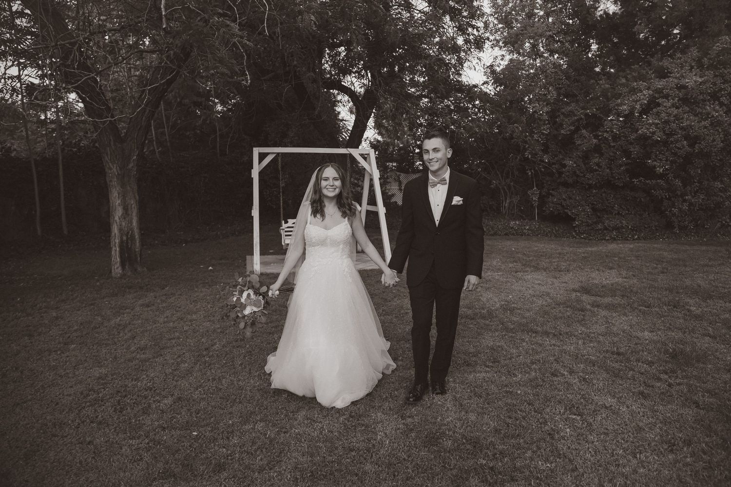 Black and white wedding portrait of a couple standing in front of a wooden arch in a garden setting.