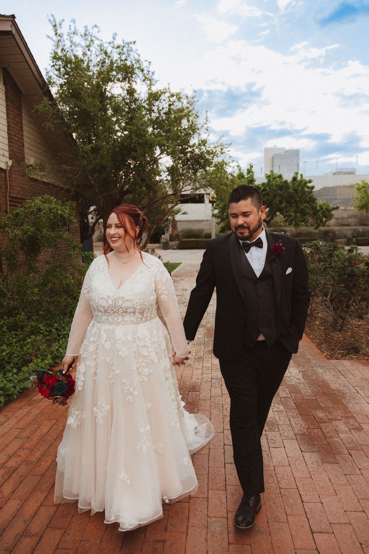 A couple holding hands walks down a brick pathway surrounded by greenery.