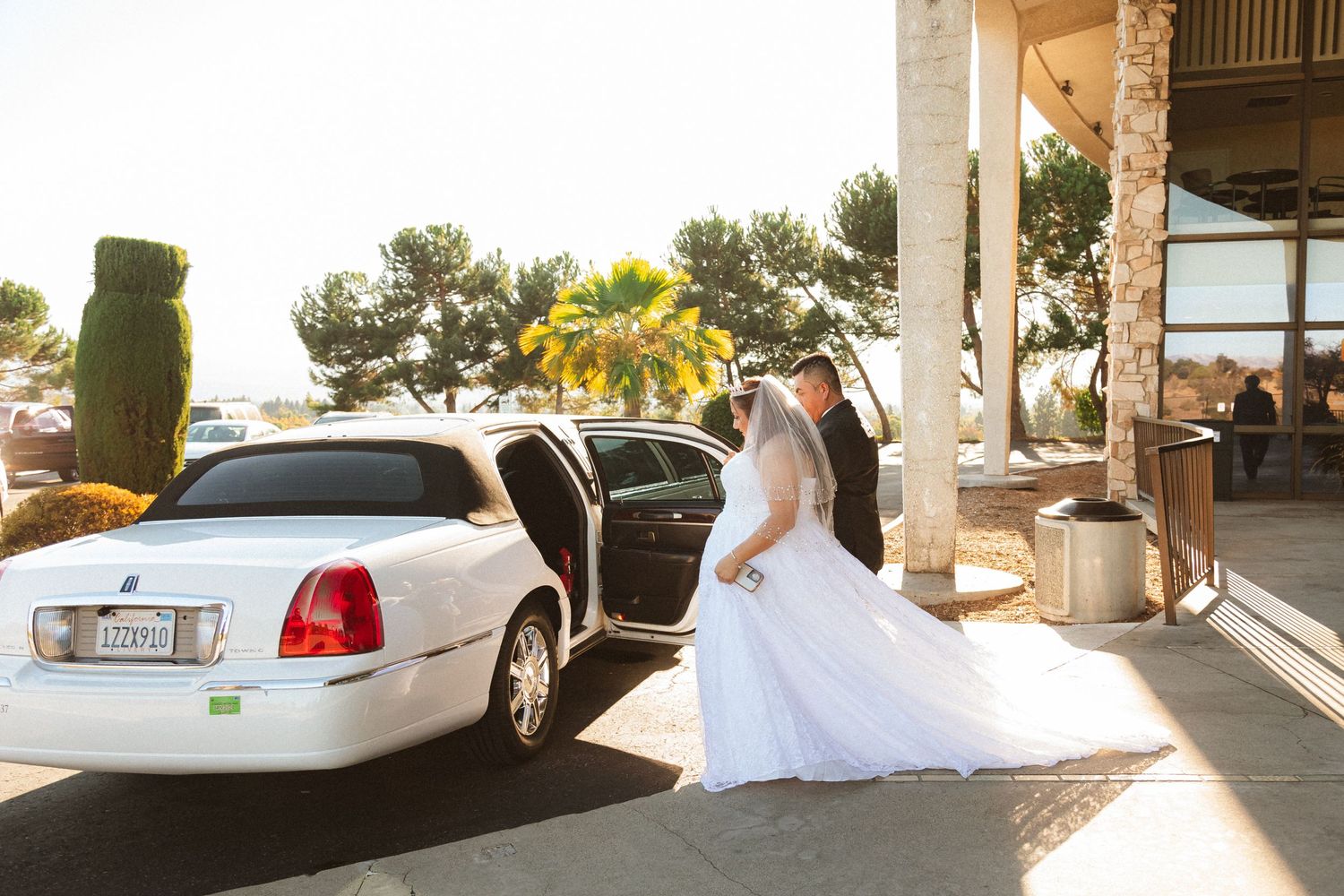 A couple in formal wedding attire walk towards a white limousine in afternoon sunlight.