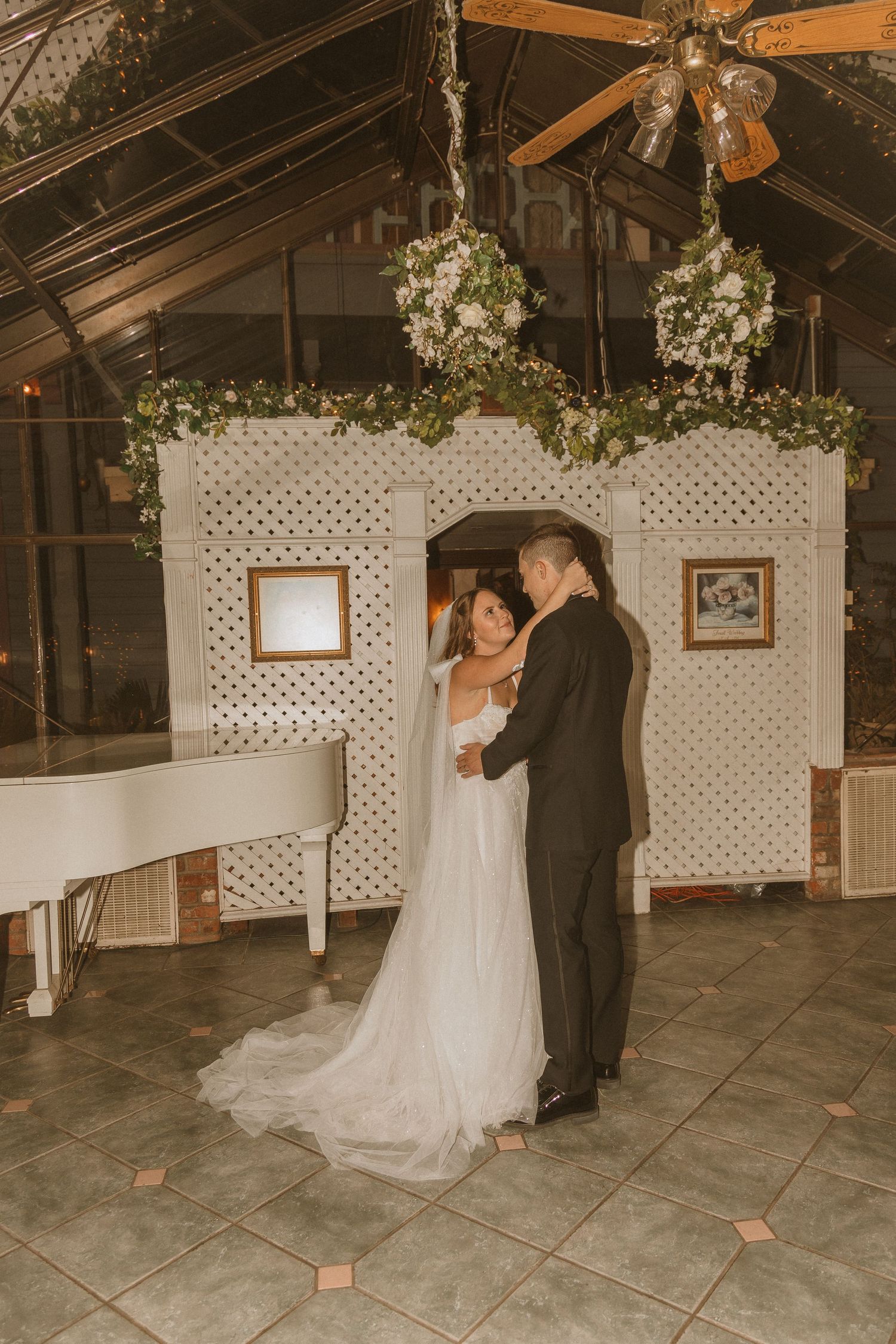 Couple dancing at wedding reception with white piano and floral decor in glass-roofed venue.