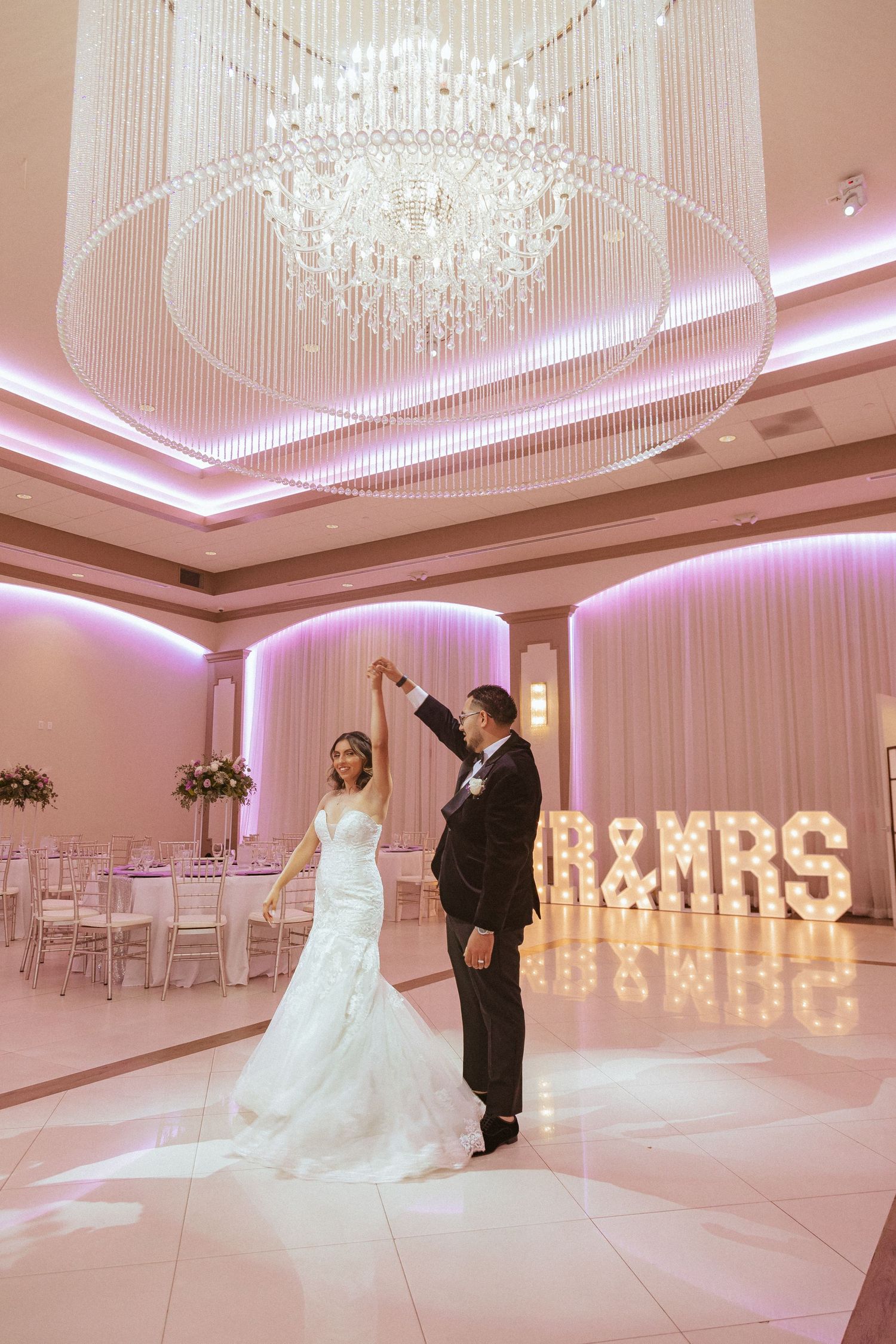 Elegant couple sharing a dance moment beneath crystal chandelier in pink-illuminated reception venue.