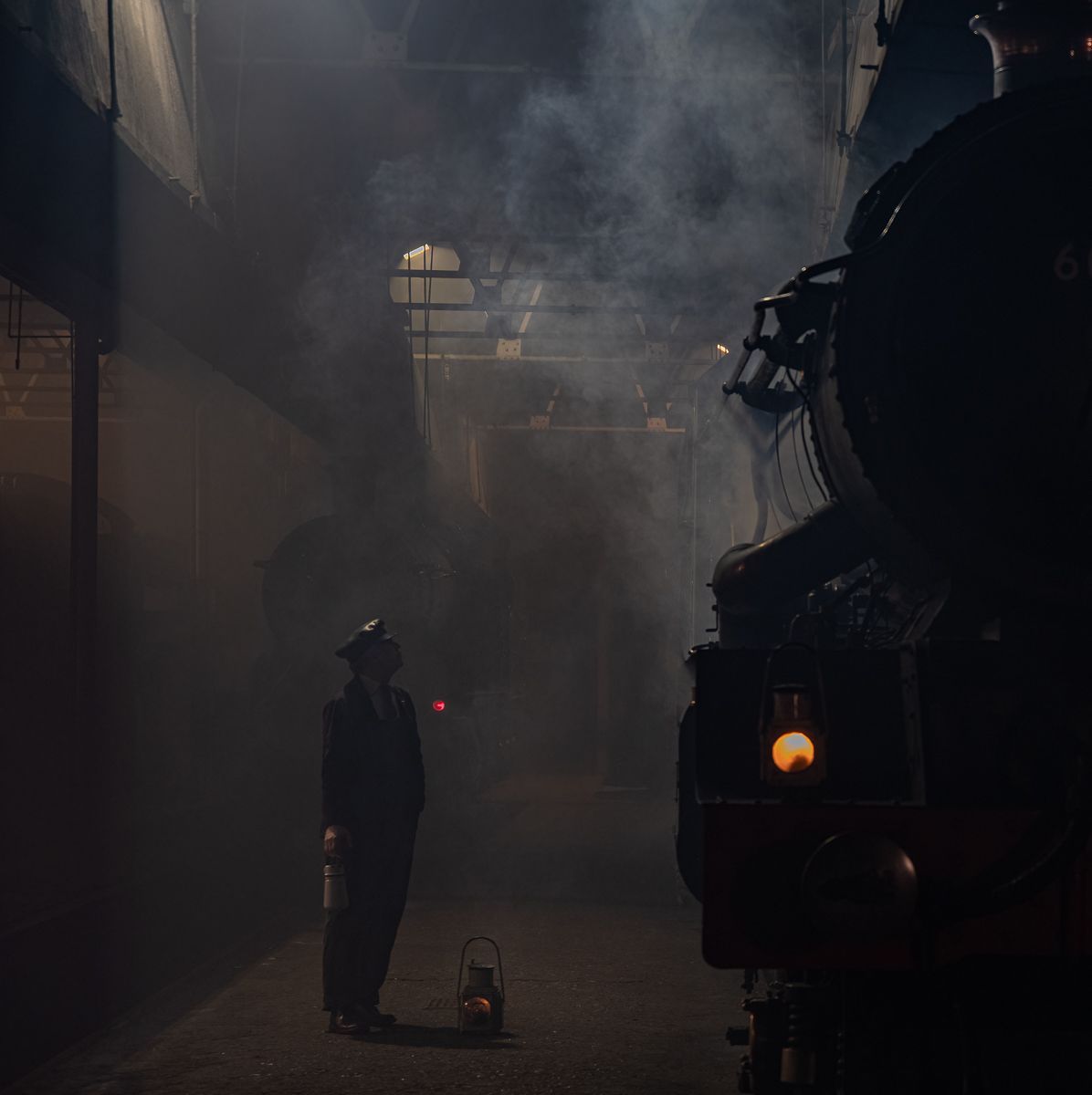 A silhouetted figure stands near a steam locomotive in atmospheric foggy darkness.