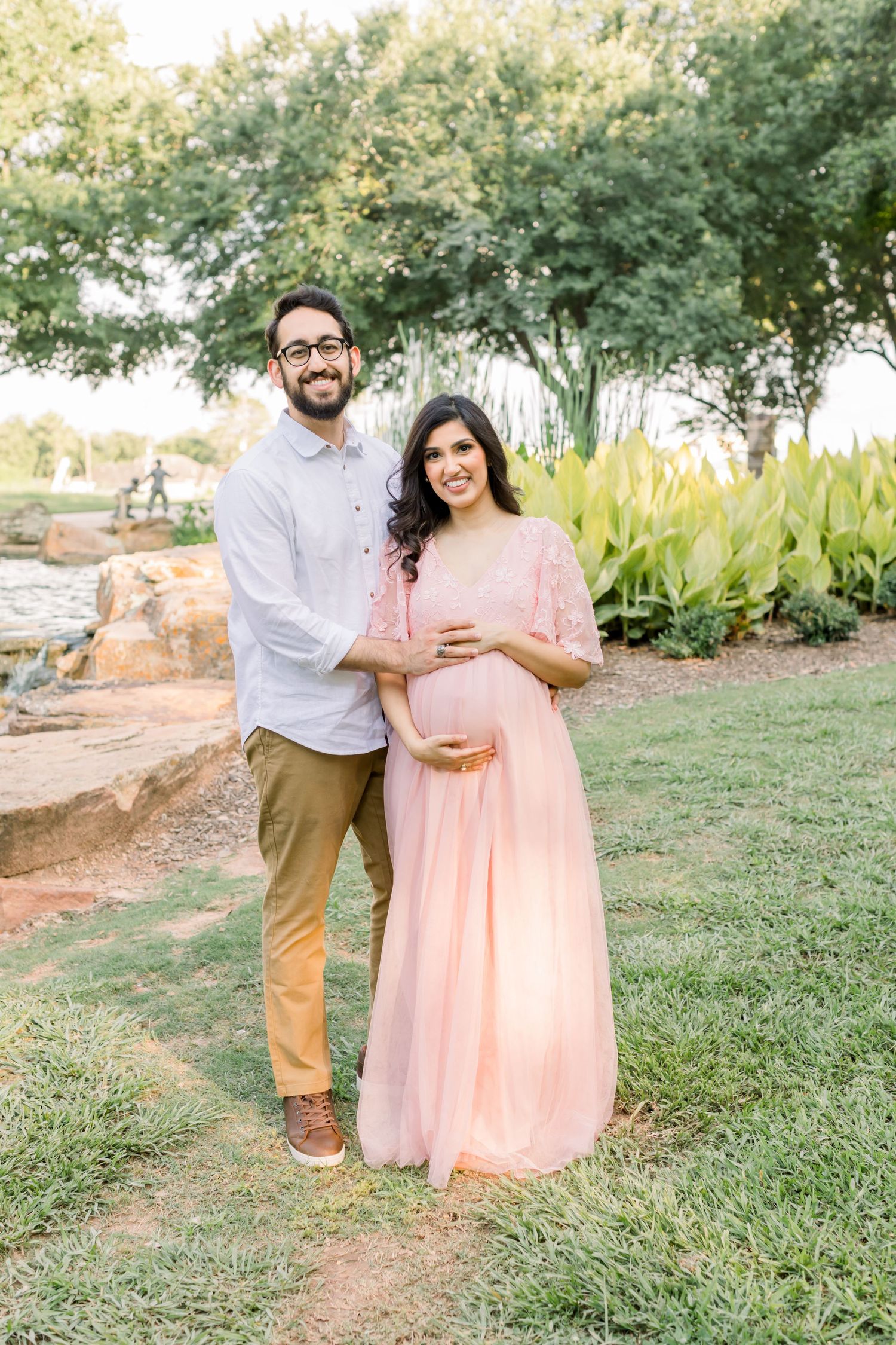 A couple poses together outdoors, with her wearing a flowing pink dress against a natural backdrop.