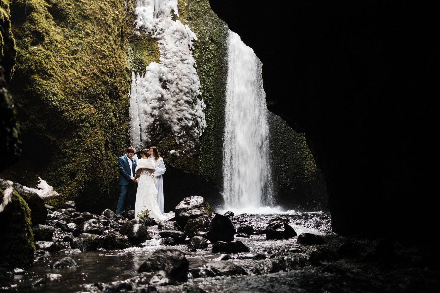 A couple poses in wedding attire beneath a dramatic waterfall in a cave setting.