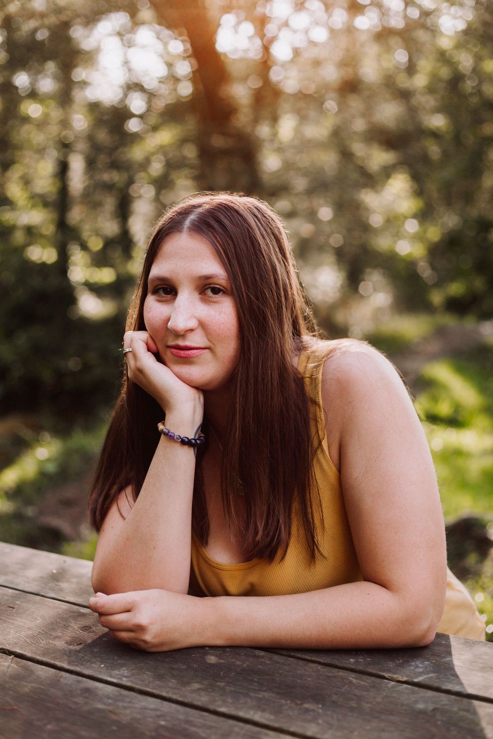 A person in a yellow top poses casually on a wooden surface in an outdoor setting with natural lighting.