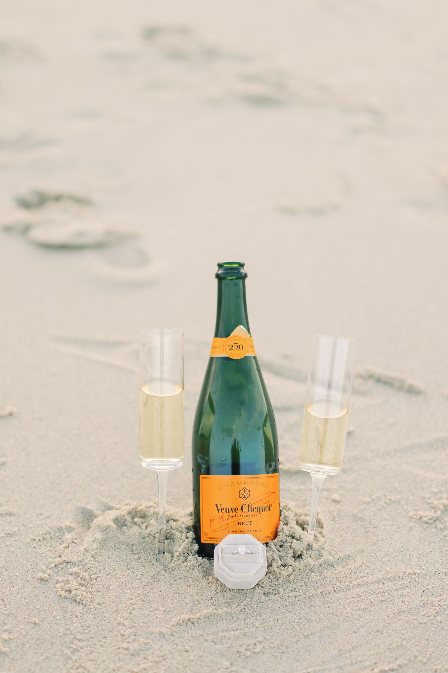 Veuve Clicquot champagne bottle with two glasses and engagement ring nestled in beach sand.