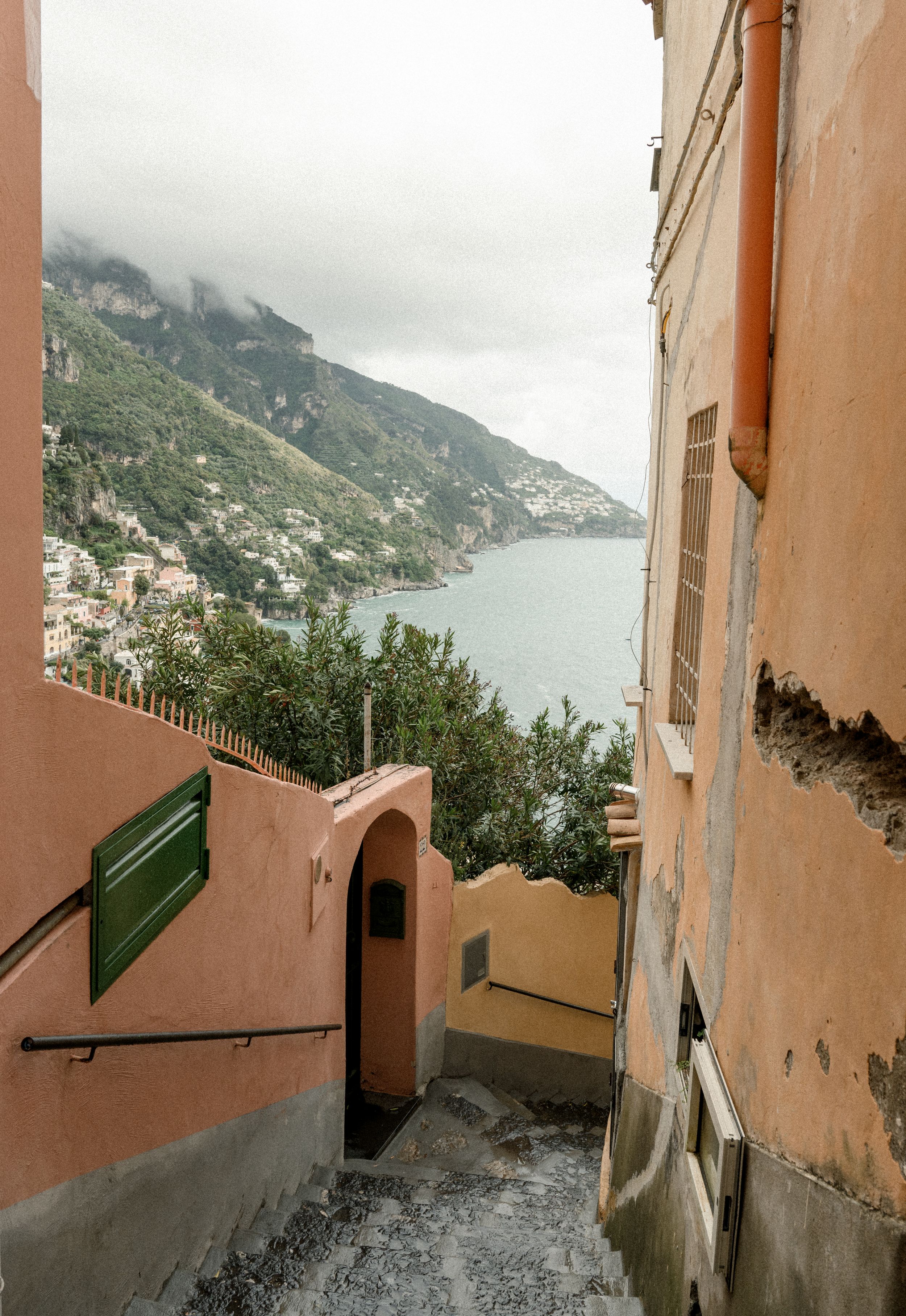 A narrow peach-colored alleyway leads down steps with Mediterranean coastal views.